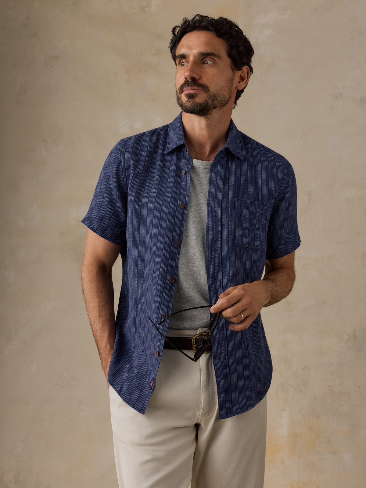 A man wearing a short-sleeve navy blue textured button-up shirt over a gray t-shirt and beige trousers, holding a pair of glasses against a textured beige background., image 1 of 6