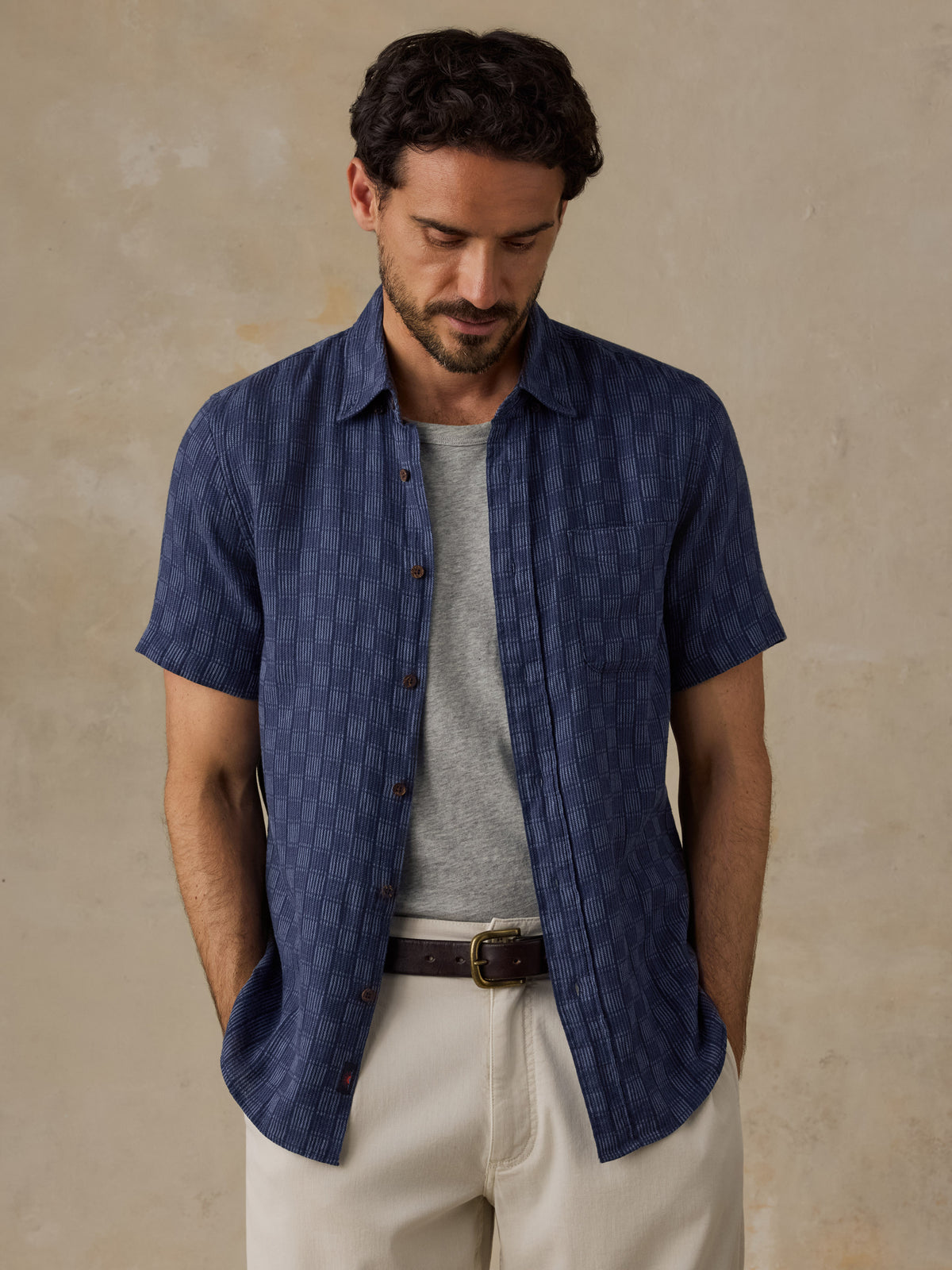 A man wearing a navy blue short-sleeve button-up shirt with a subtle textured pattern, layered over a gray t-shirt and beige trousers, standing against a textured beige wall., image 6 of 6