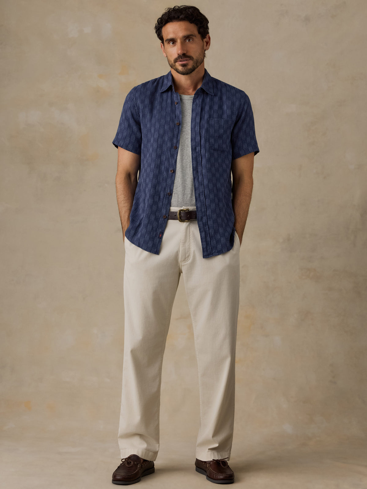 A man stands against a textured beige studio backdrop wearing a navy short-sleeve button-up shirt with a subtle geometric pattern, layered over a light gray t-shirt, cream-colored trousers, and dark brown boat shoes., image 5 of 6
