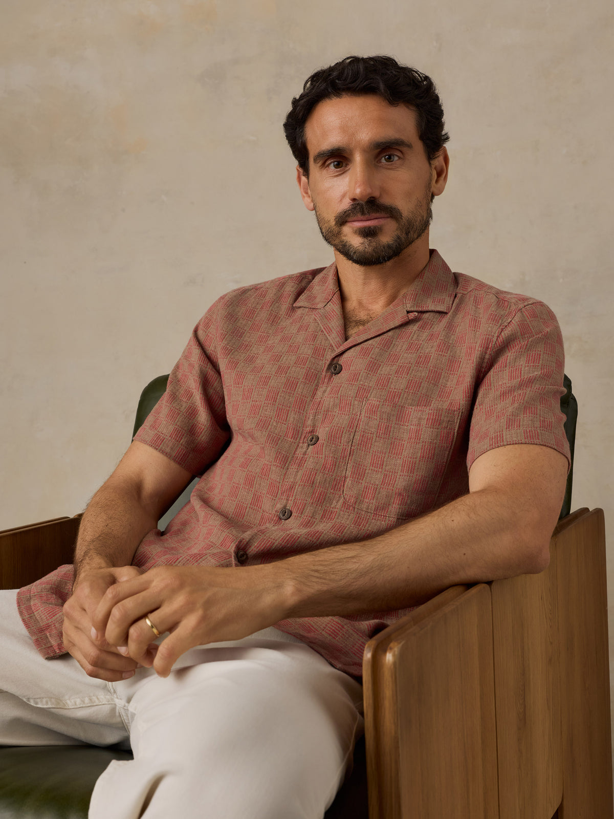A man wearing a short-sleeve button-up shirt with a muted red and beige geometric pattern, paired with white trousers, seated in a wooden armchair against a textured beige wall., image 6 of 7