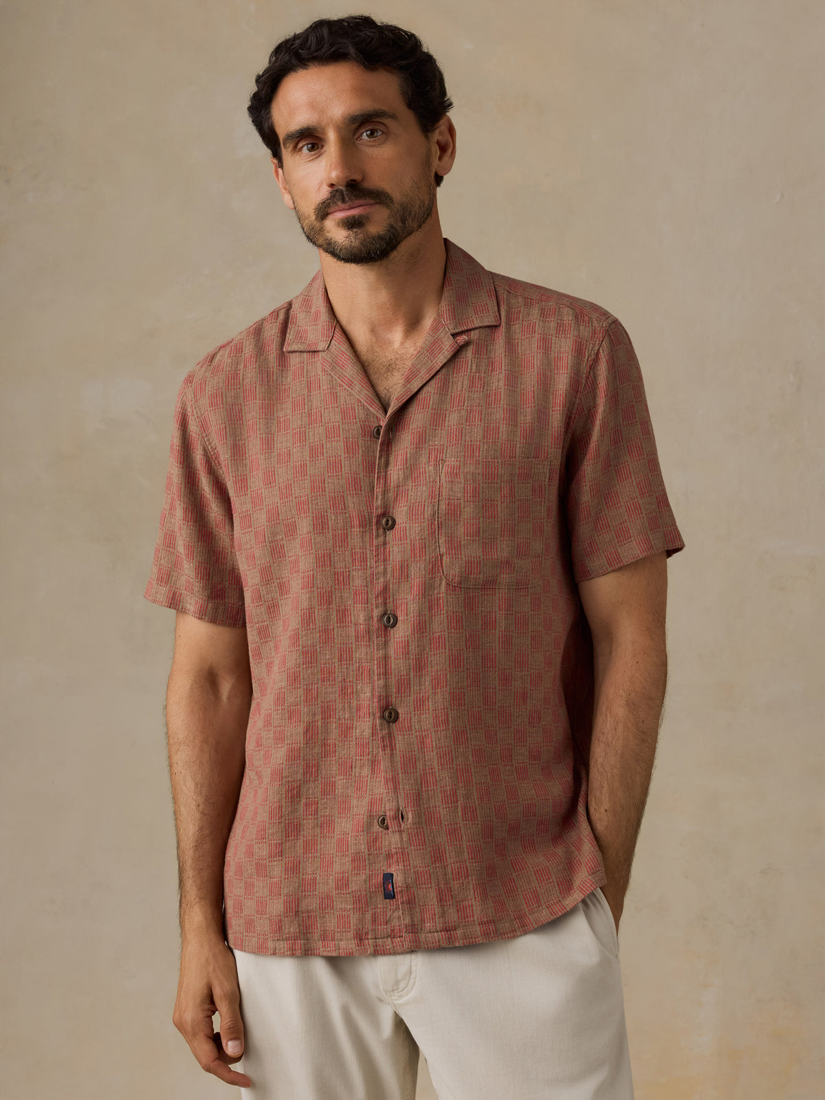A man wearing a short-sleeve button-up shirt with a muted red and brown geometric pattern, paired with off-white trousers, standing against a textured beige background., image 2 of 7