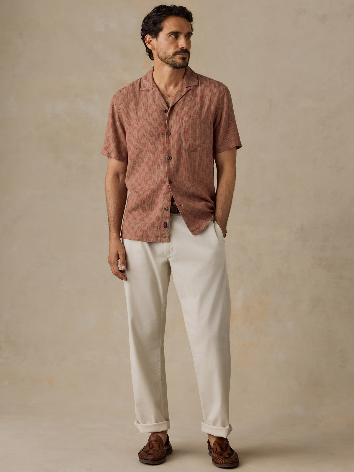 A man wearing a short-sleeve button-up shirt in a dusty rose-brown color with a subtle geometric pattern, paired with off-white trousers and brown shoes, standing against a textured beige background., image 5 of 7