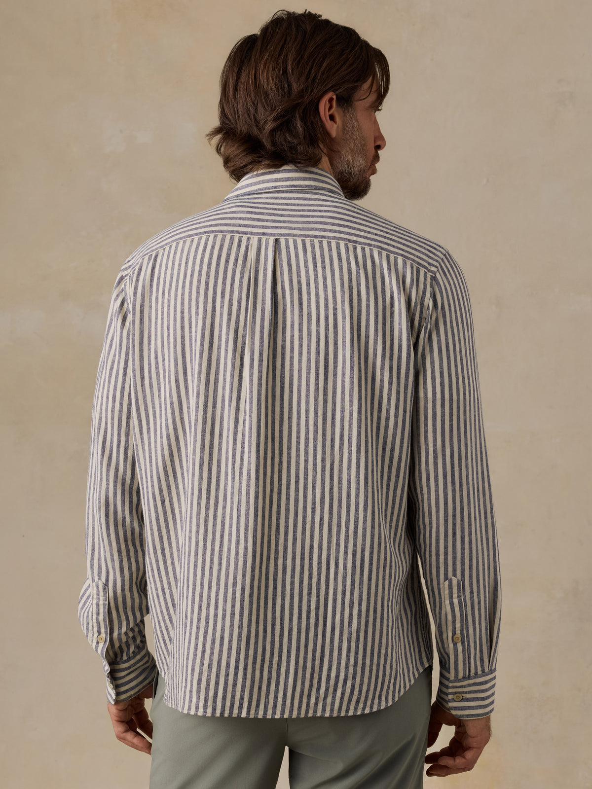 A man viewed from behind wearing a long-sleeve button-up shirt with vertical gray and white stripes, paired with muted green shorts, set against a textured beige background., image 5 of 6