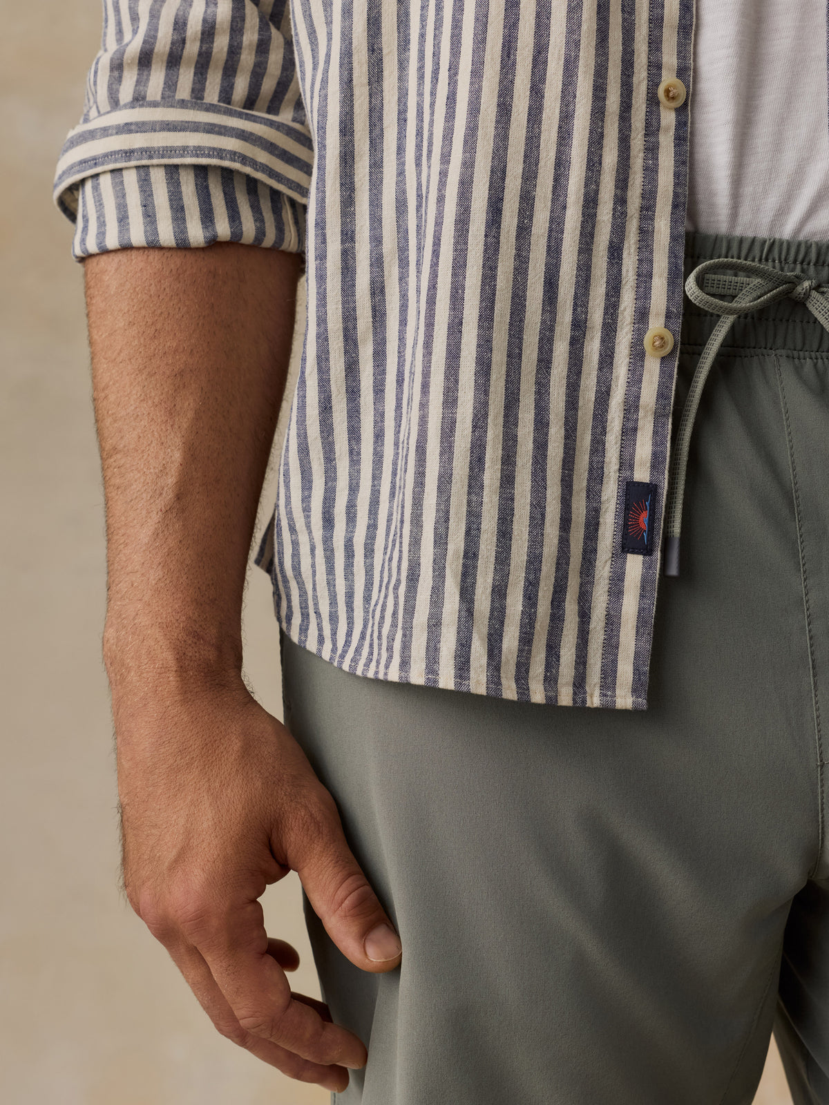 Close-up of a man wearing a blue and white vertically striped button-up shirt with rolled sleeves, paired with gray drawstring trousers., image 3 of 6