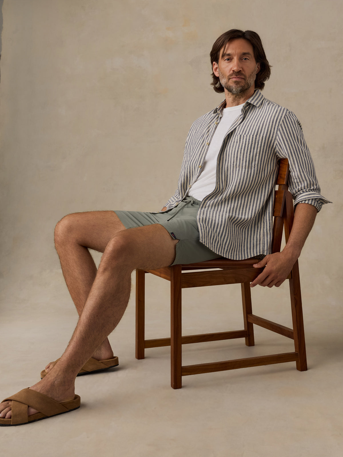 A man sits on a wooden chair wearing a white t-shirt, a striped button-down shirt, sage green shorts, and brown slide sandals against a textured beige studio backdrop., image 4 of 6