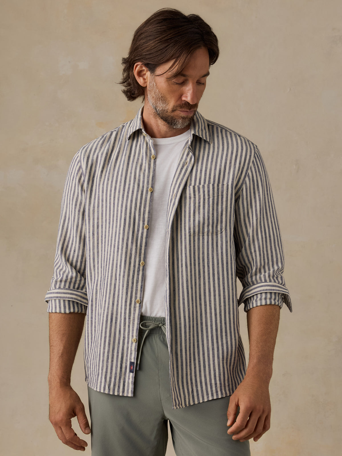 A man wearing a long-sleeve button-up shirt with vertical gray and white stripes over a white t-shirt, paired with olive green pants, set against a textured beige background., image 2 of 6