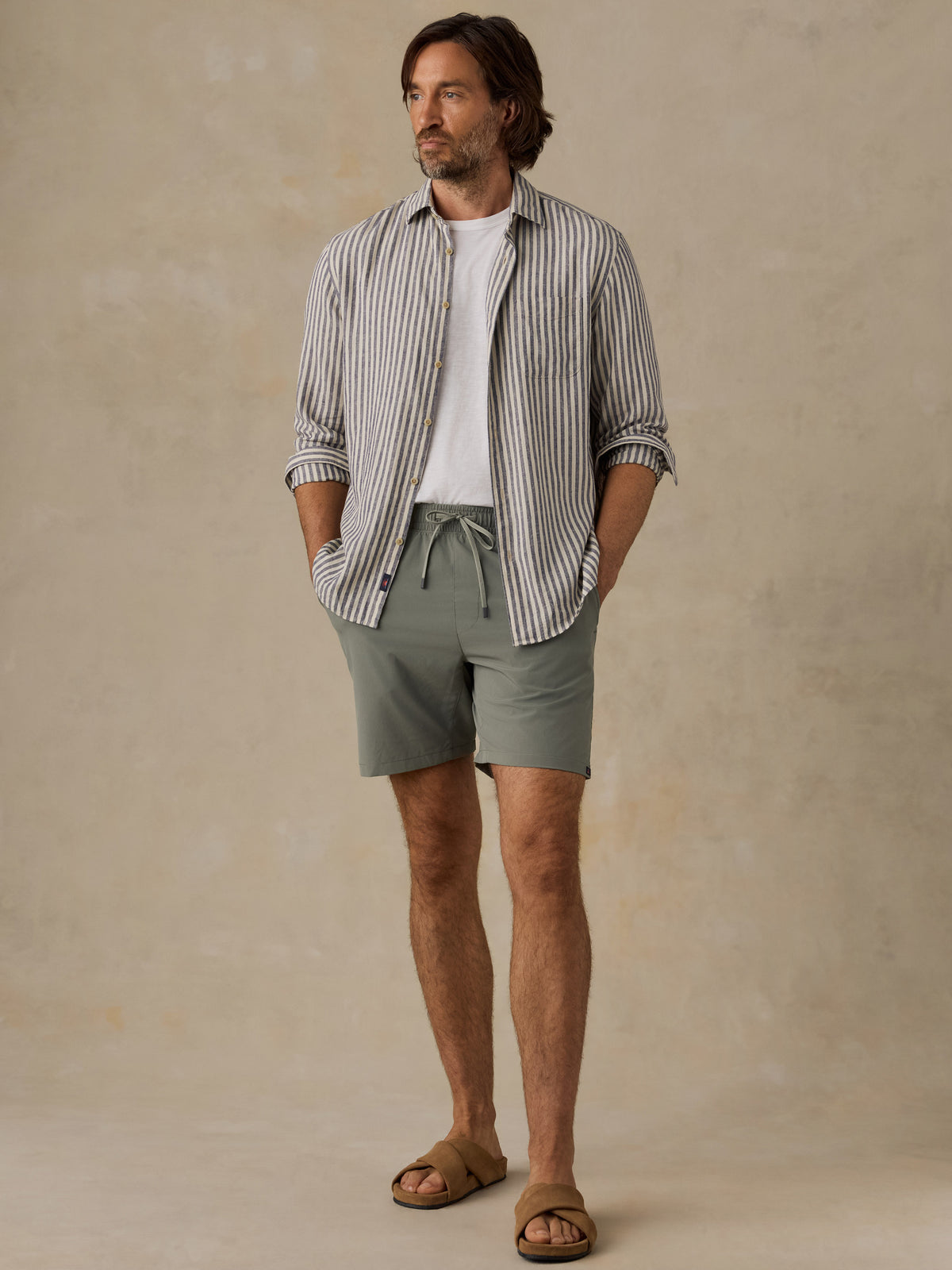 A man stands against a warm beige textured studio backdrop wearing a striped button-up shirt over a white tee, sage green drawstring shorts, and brown slide sandals., image 6 of 6