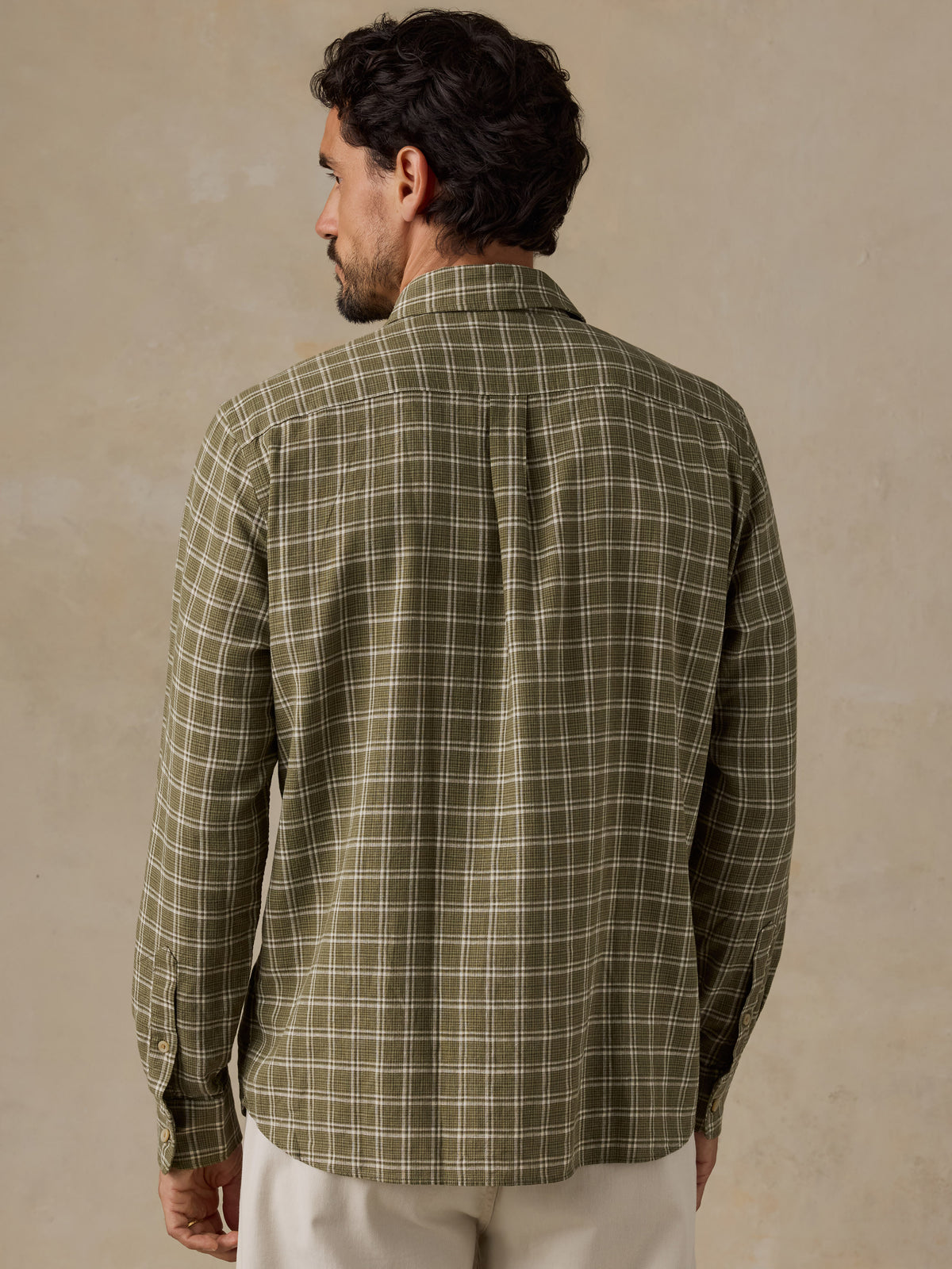 A man shown from the back wearing a long-sleeve plaid button-up shirt in olive green and beige, paired with off-white trousers, against a textured beige background., image 3 of 6