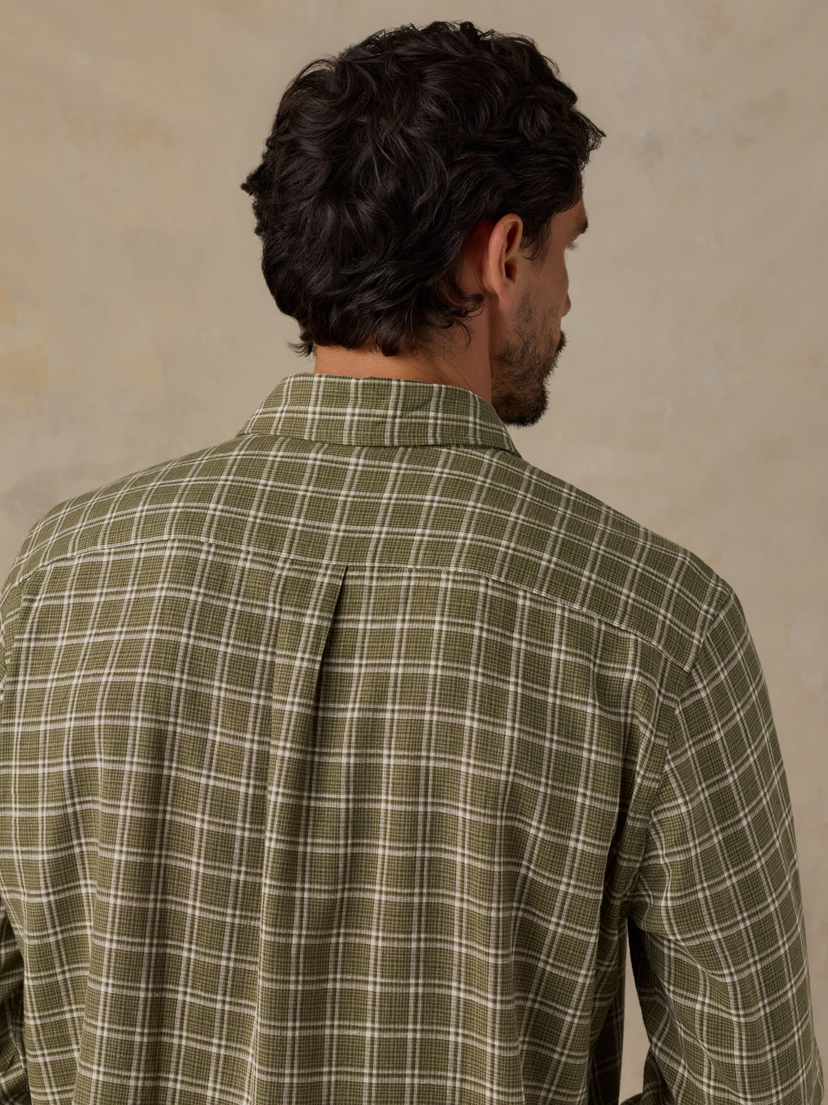 A man viewed from behind wearing a long-sleeve plaid button-down shirt in olive green and white, set against a textured beige background., image 6 of 6