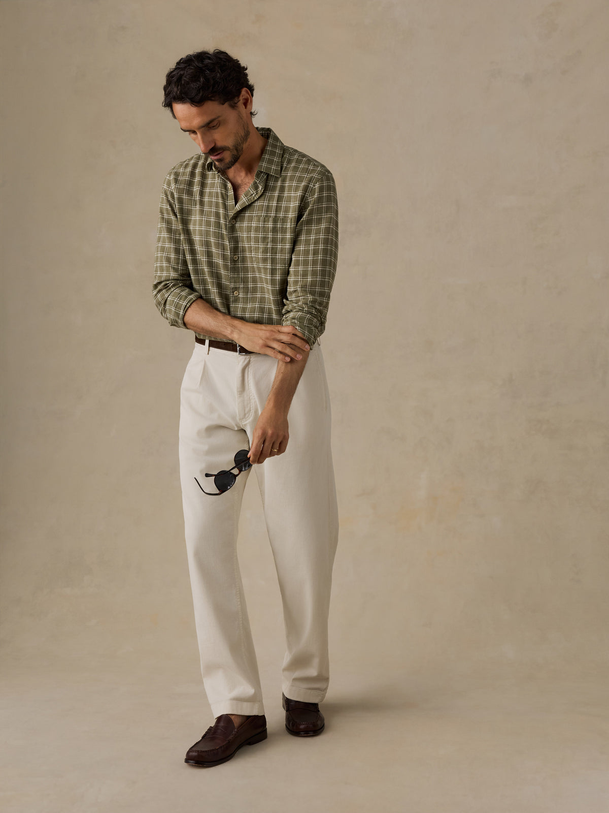 A man wearing a green and beige plaid button-down shirt, off-white trousers, and dark brown loafers, standing against a textured beige studio backdrop., image 5 of 6