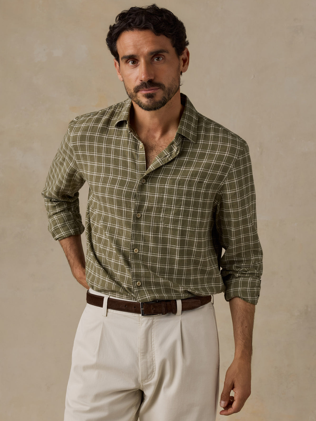 A man wearing a long-sleeve button-up shirt with an olive green and brown plaid pattern, paired with off-white trousers and a brown belt, standing against a textured beige background., image 1 of 6