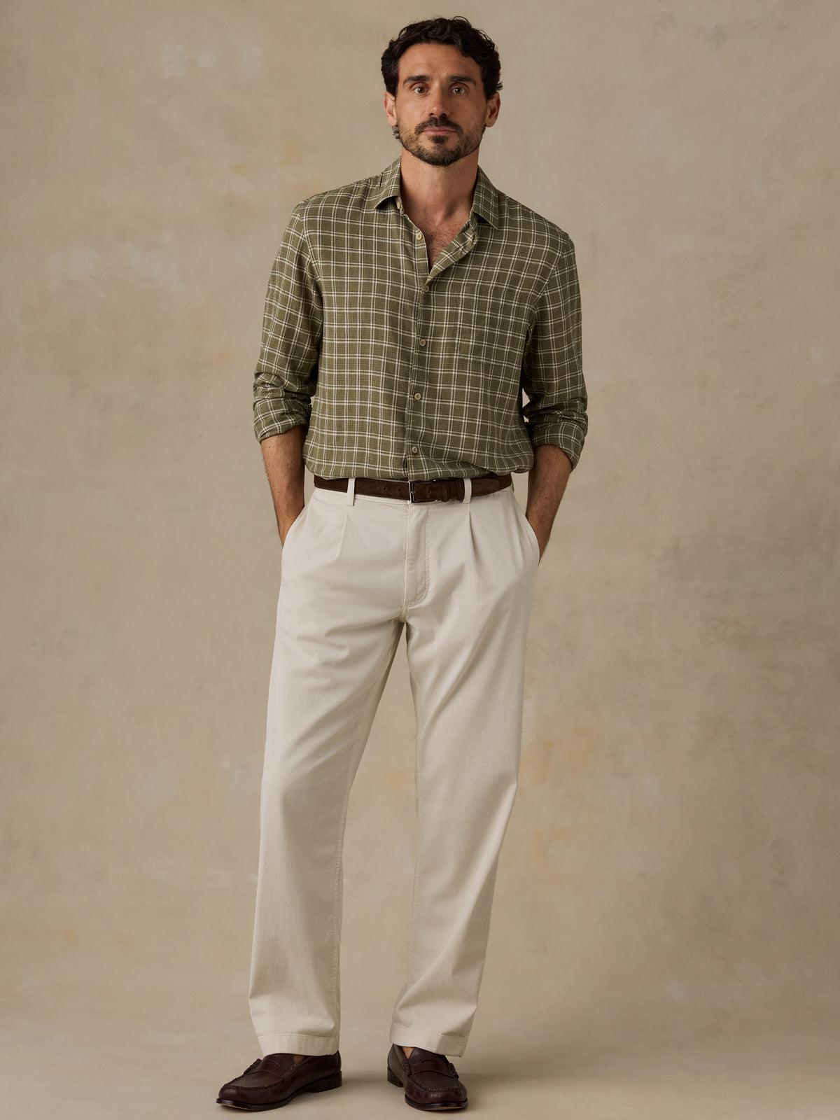 A man wearing a green and beige plaid button-down shirt, off-white trousers, and dark brown loafers, standing against a textured beige studio backdrop., image 2 of 6