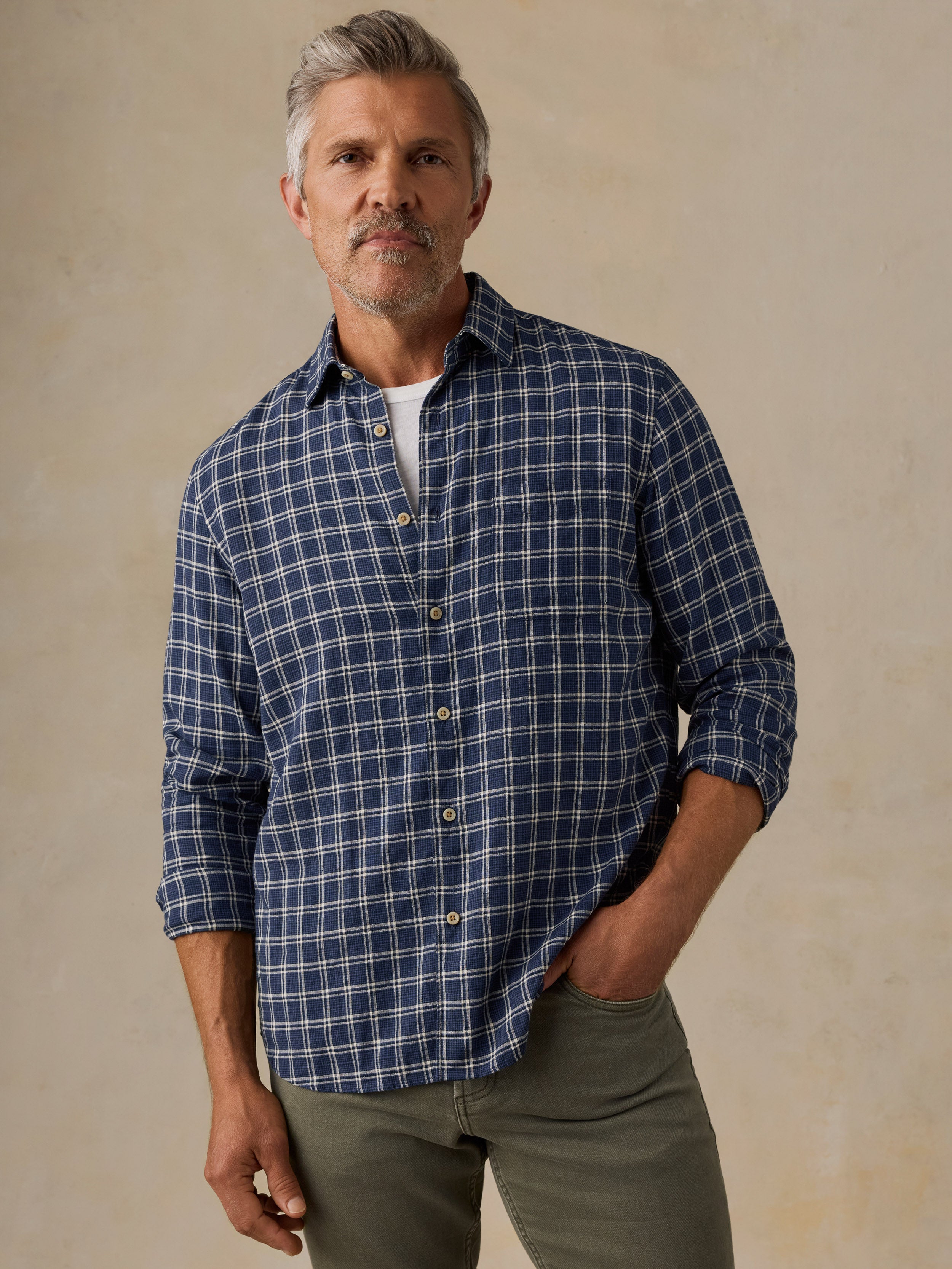 A man wearing a navy blue and white plaid button-down shirt with olive green pants, standing against a textured beige background.