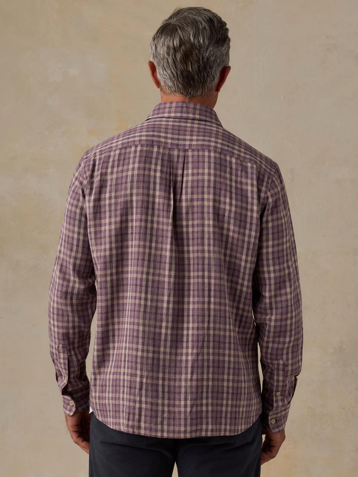 A man viewed from behind wearing a long-sleeve plaid button-down shirt in a muted purple and beige pattern, paired with dark trousers, against a textured beige background., image 3 of 6