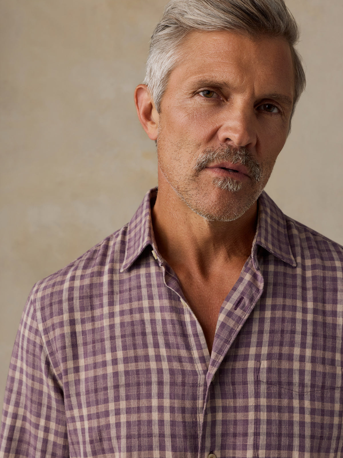 A close-up portrait of a man with gray hair and a mustache wearing a purple and beige plaid button-down shirt against a textured beige background., image 2 of 6