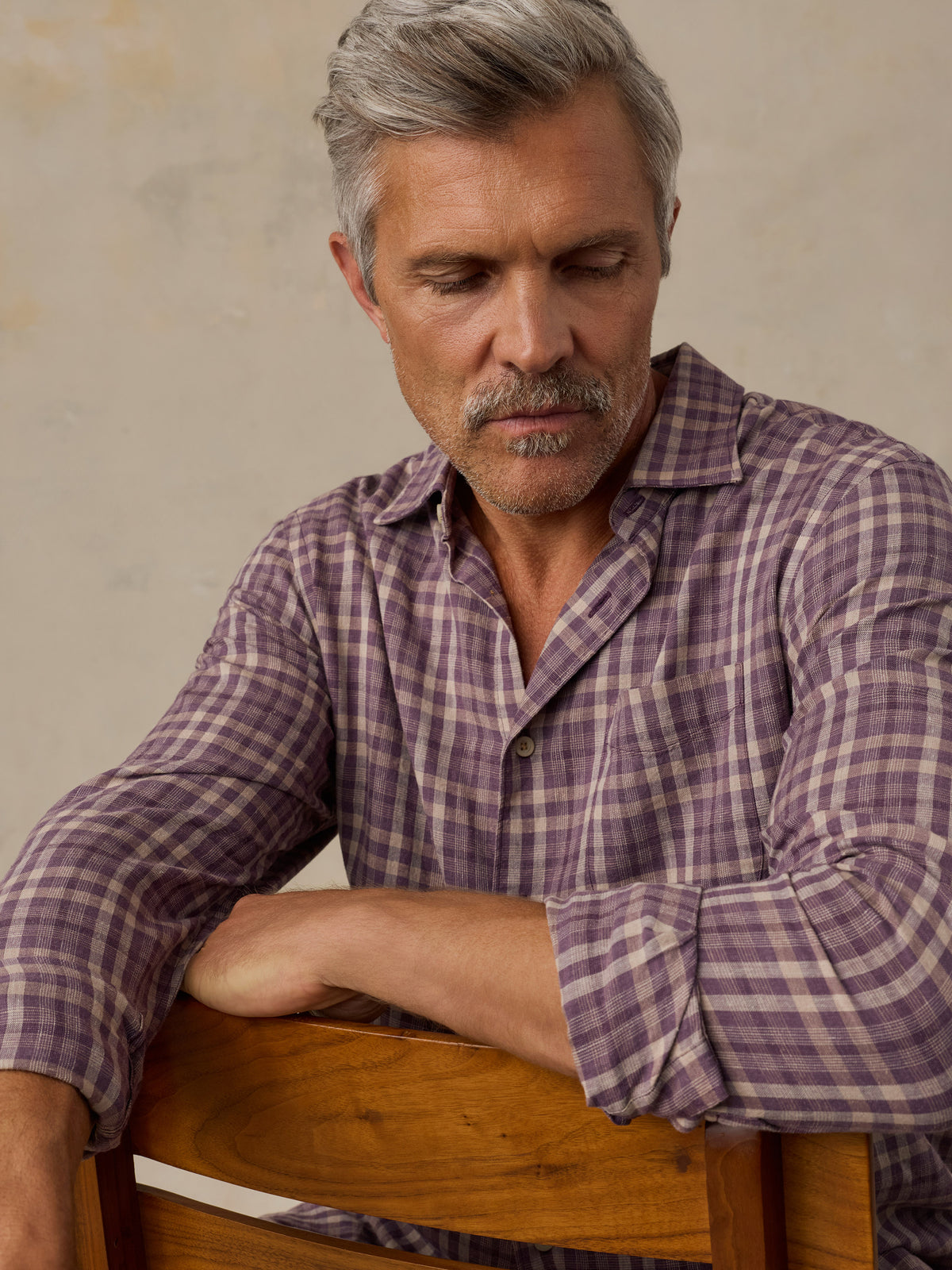A man with gray hair and a mustache wearing a purple and beige plaid button-down shirt, leaning on a wooden chair against a textured beige wall., image 6 of 6