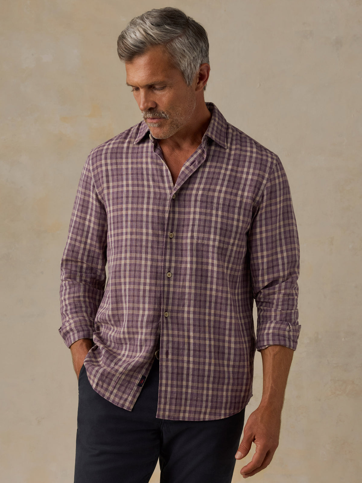 A man wearing a long-sleeve button-down plaid shirt in a dusty purple and brown pattern, paired with dark trousers, standing against a textured beige background., image 1 of 6