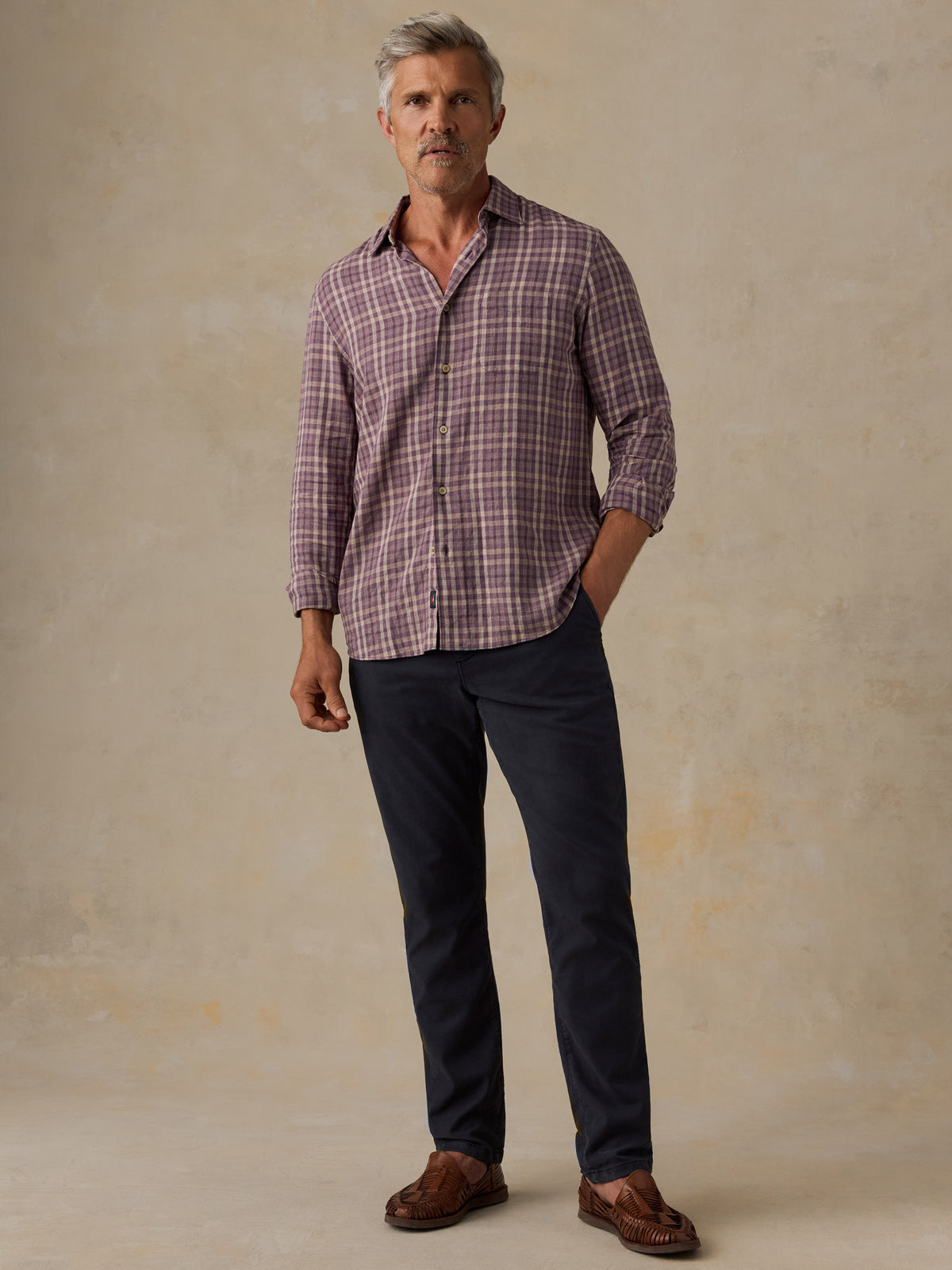 A man wearing a long-sleeve plaid button-down shirt in muted purple and white, paired with dark navy trousers and brown loafers, standing against a textured beige studio backdrop., image 5 of 6