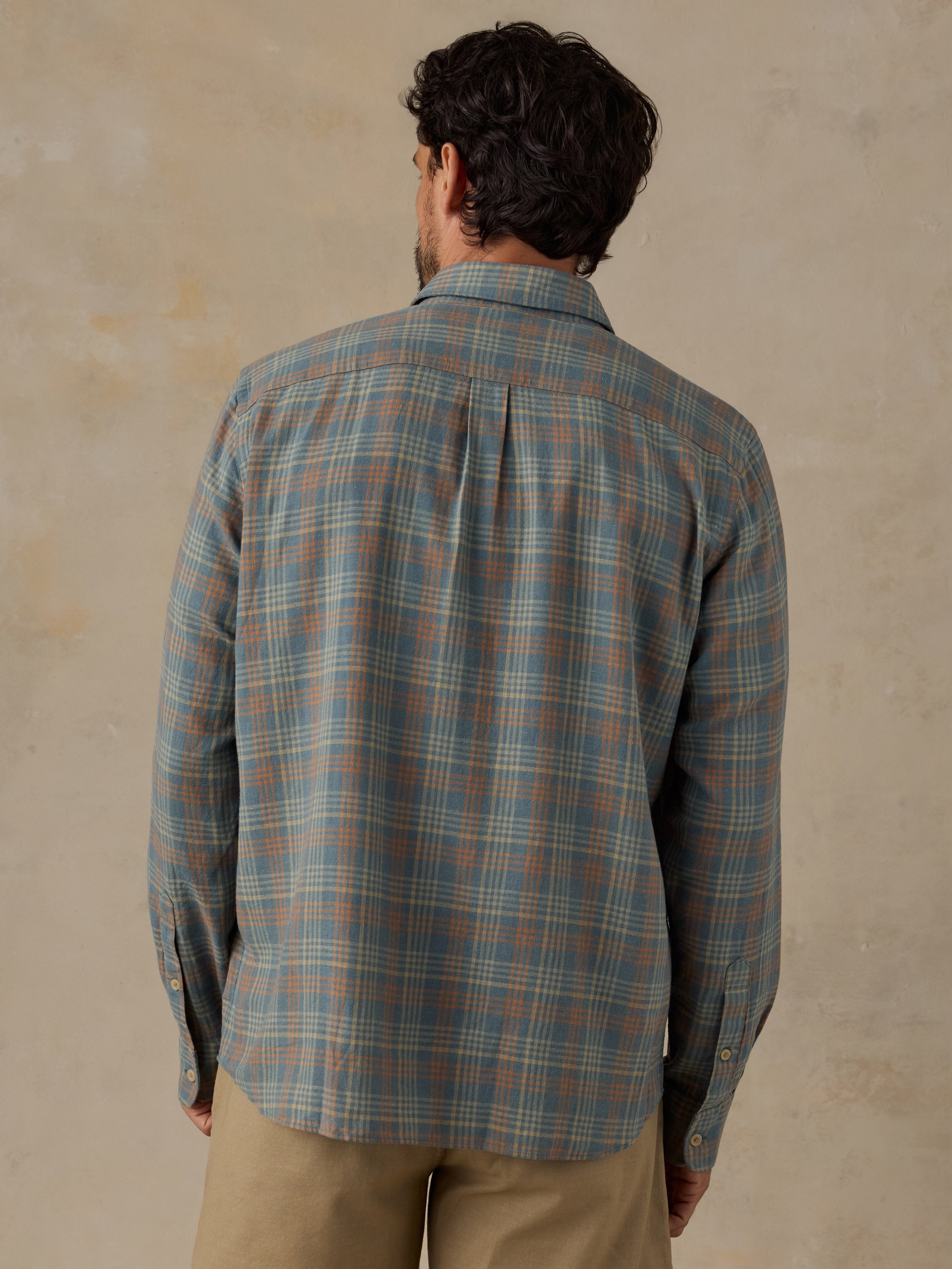 A man viewed from behind wearing a long-sleeve plaid button-up shirt in muted blue-gray, beige, and brown tones, paired with khaki pants, against a textured beige wall.