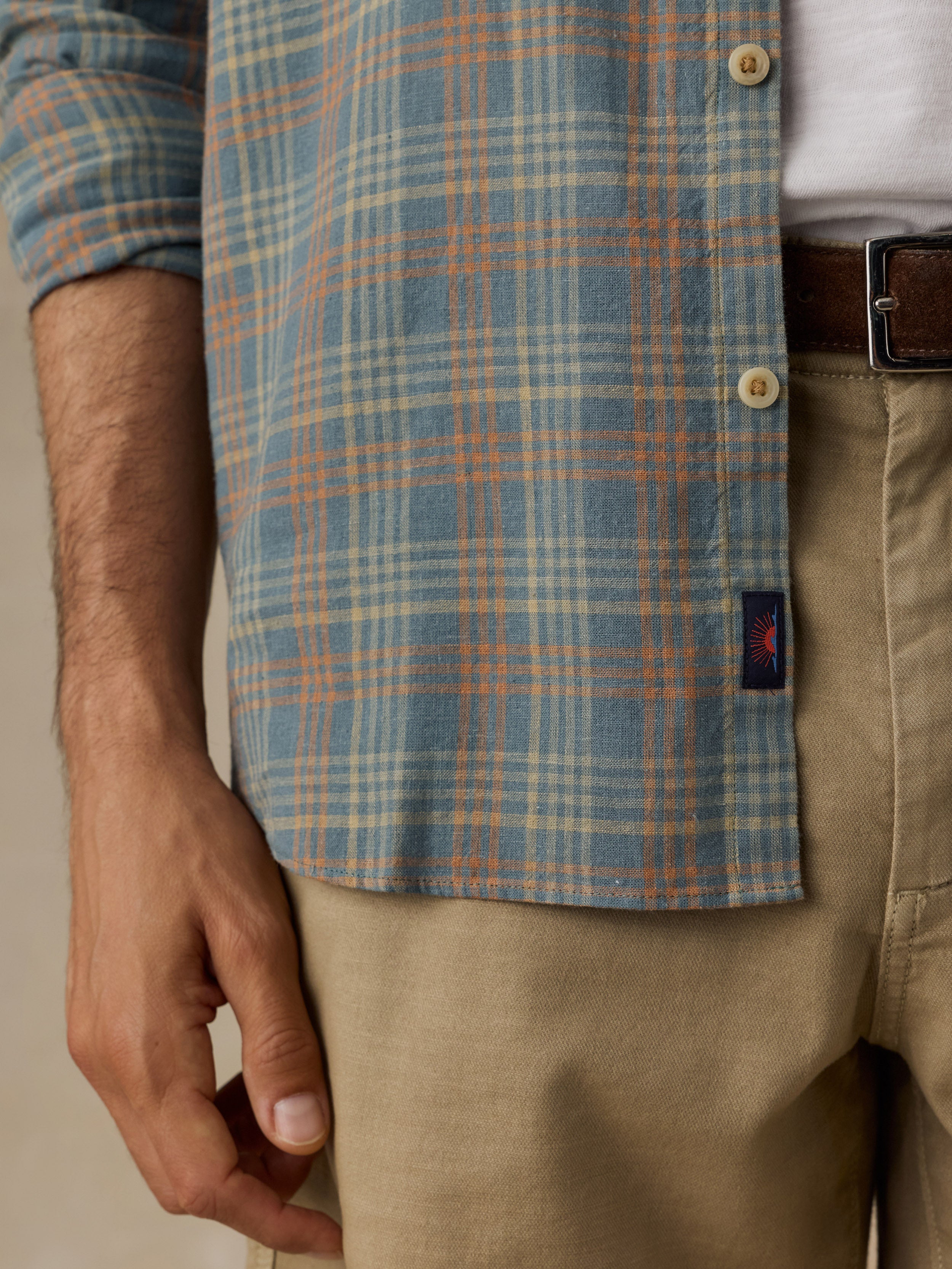Close-up of a man wearing a blue and orange plaid button-down shirt over a white undershirt, paired with khaki pants and a brown belt.