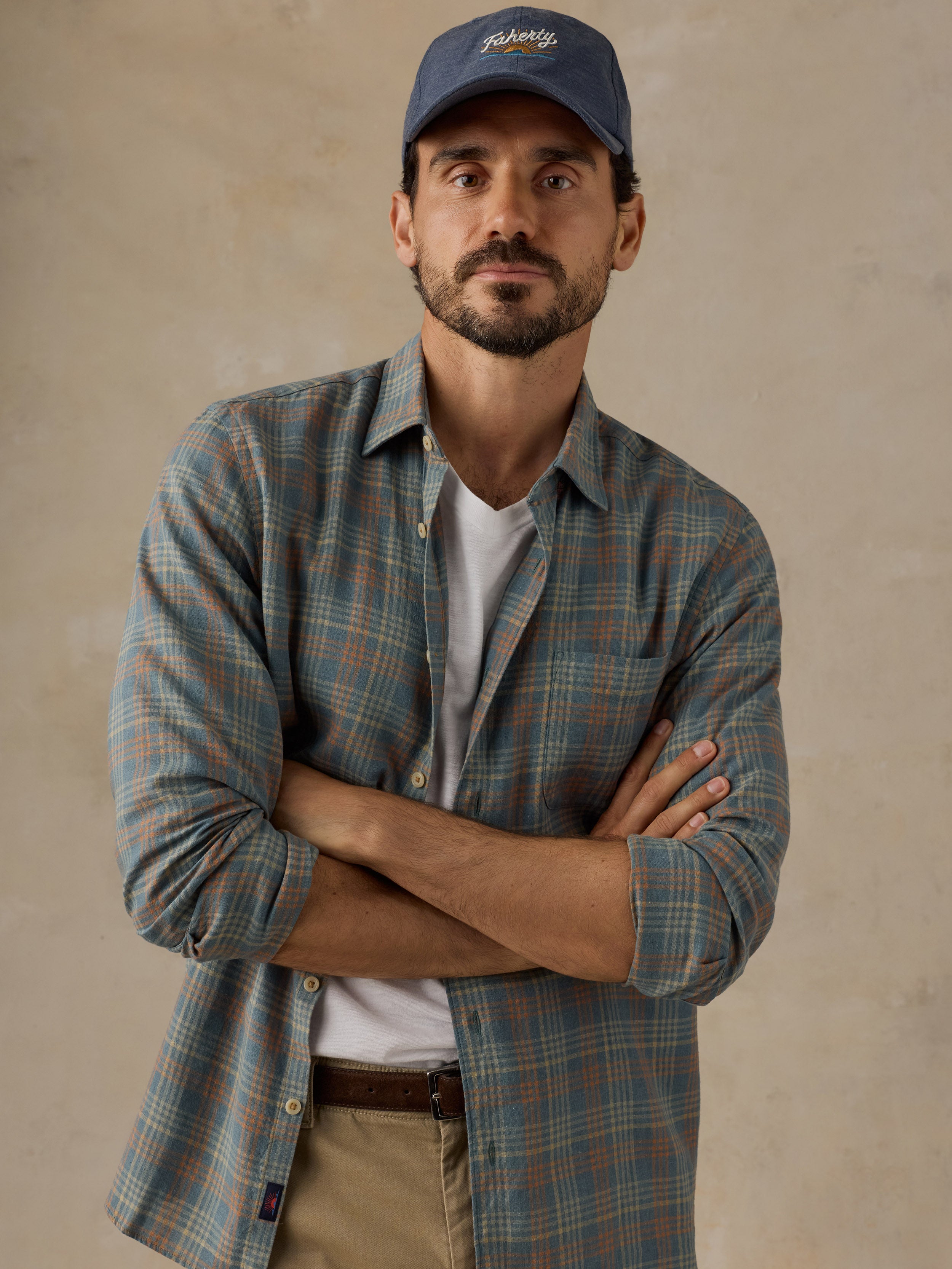A man wearing a blue and green plaid button-down shirt over a white t-shirt, khaki pants, and a blue baseball cap with 'Faherty' embroidered on the front, standing with arms crossed against a textured beige wall.