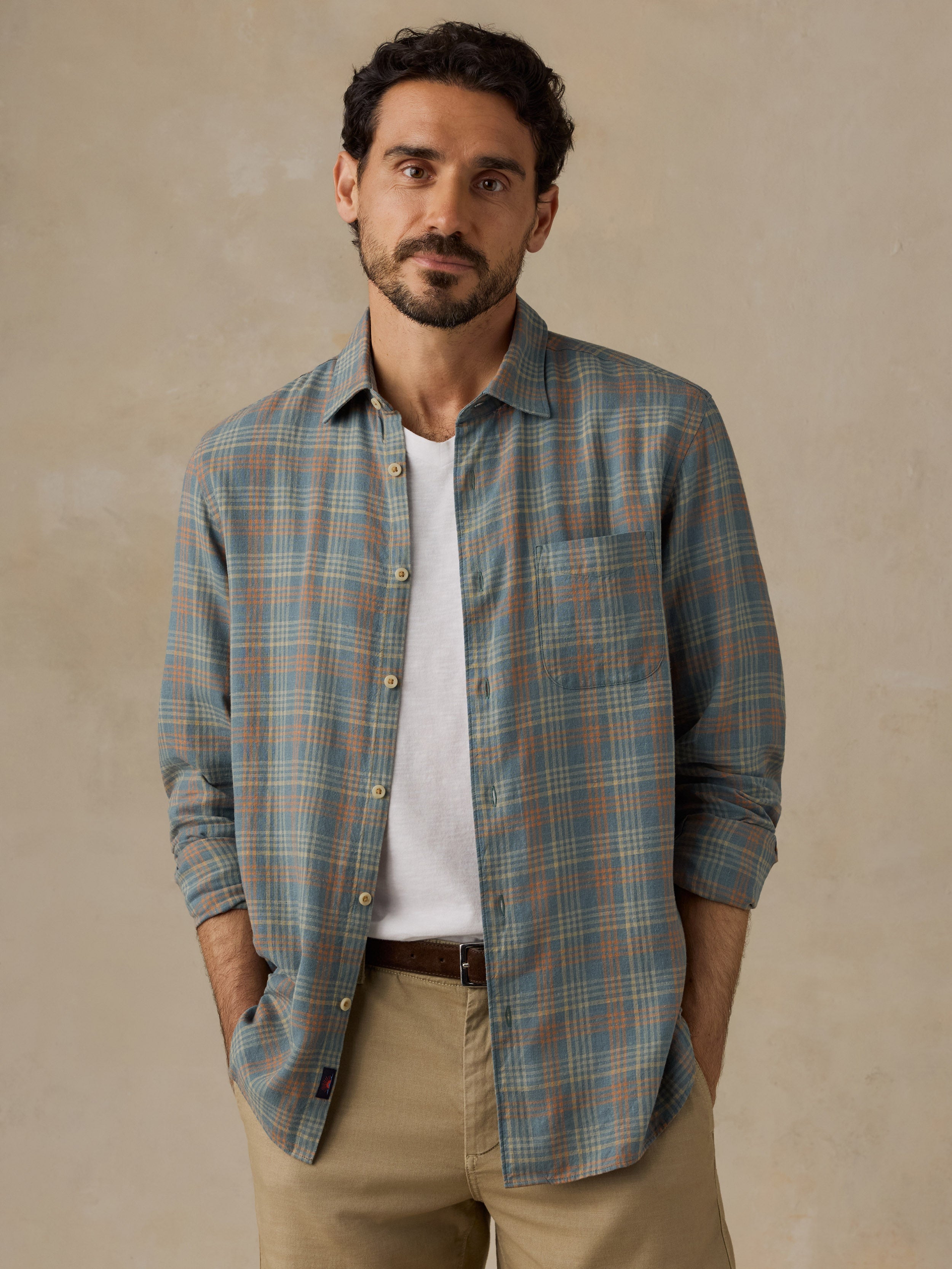 A man wearing a muted blue-green plaid button-down shirt over a white t-shirt and khaki pants, standing against a textured beige wall.