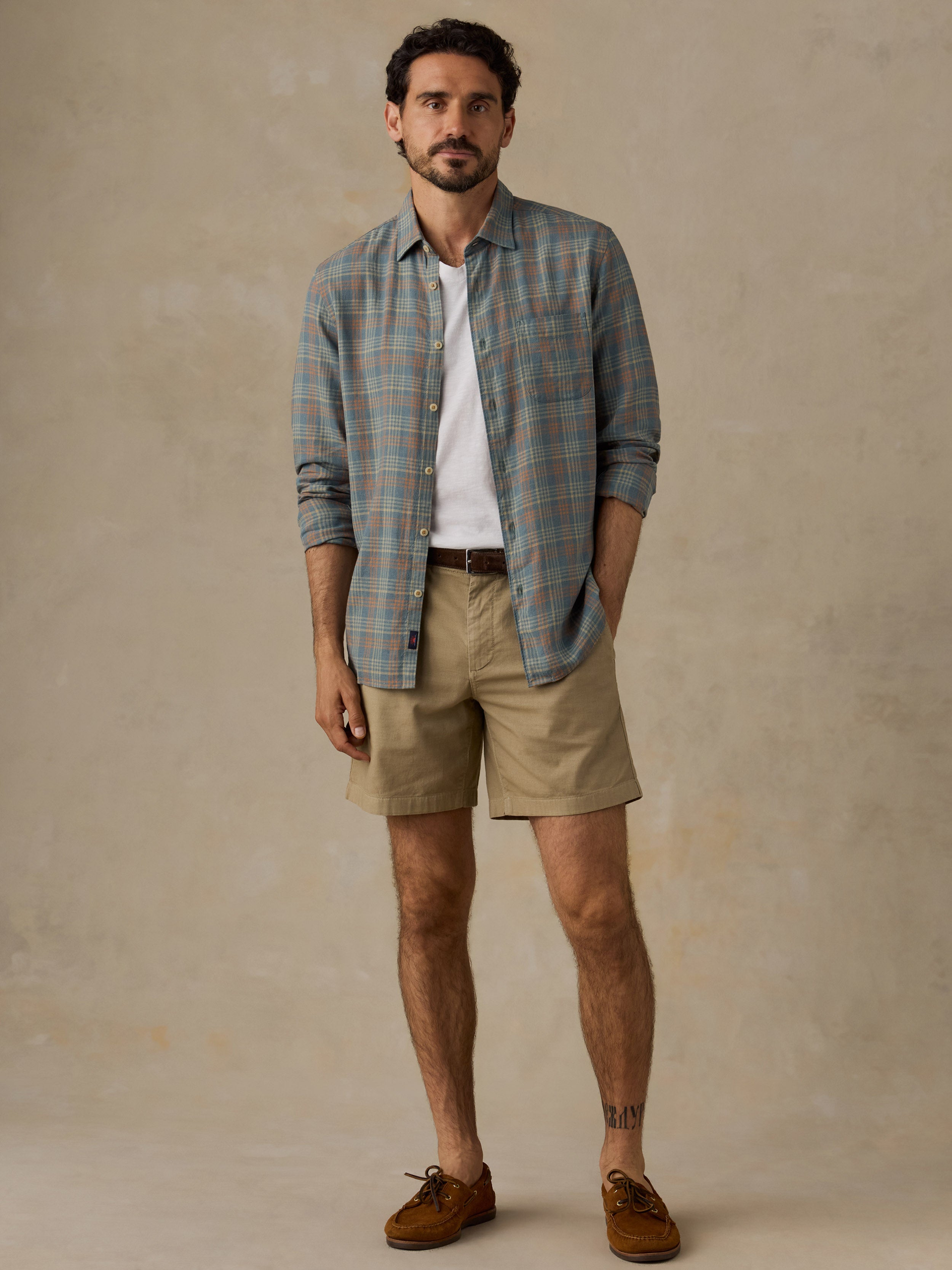 A man modeling a long-sleeve plaid button-down shirt in muted blue and green tones over a white t-shirt, paired with khaki shorts and brown suede shoes, against a textured beige studio backdrop.