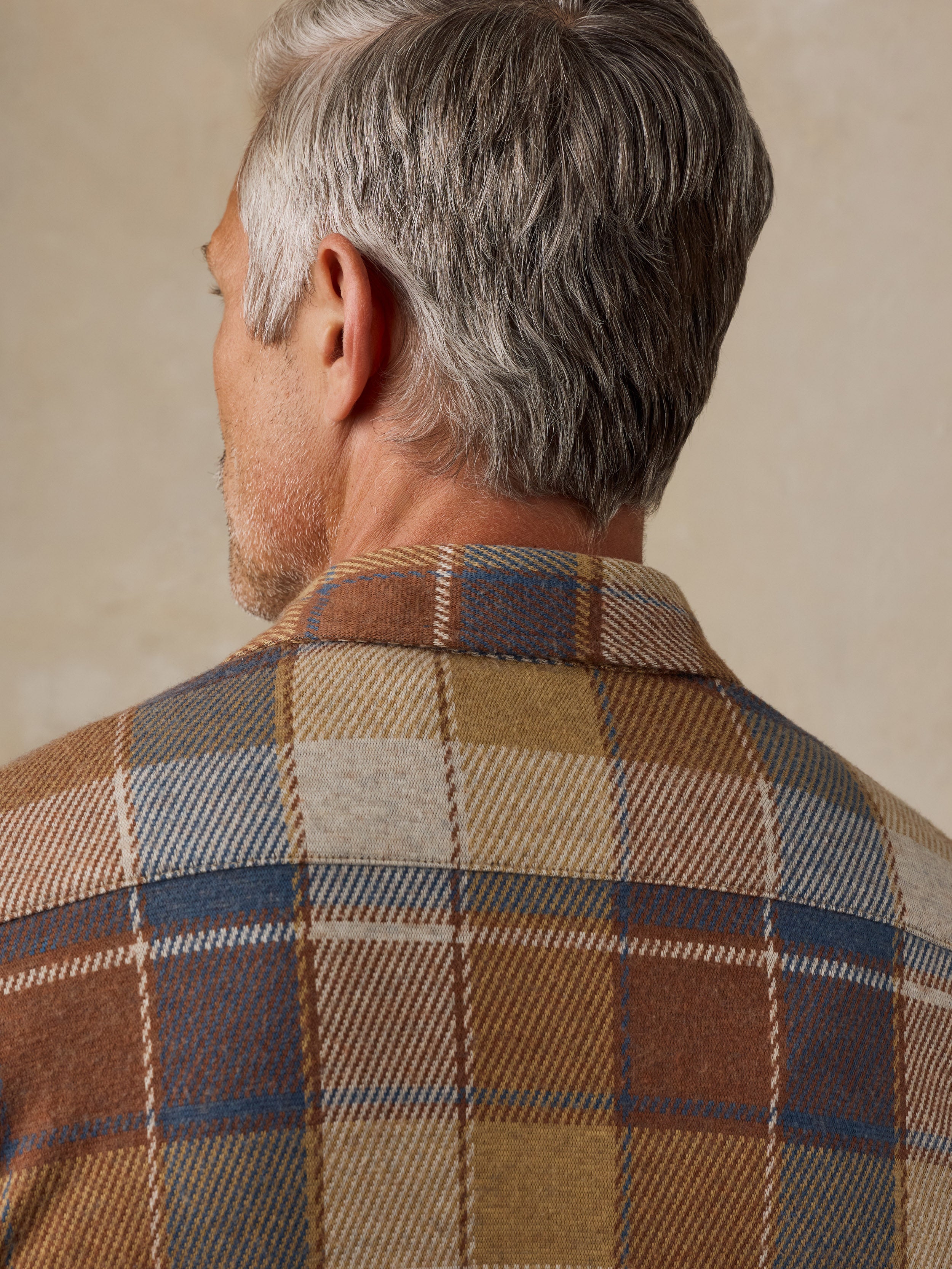 Close-up back view of a man wearing a brown, beige, and blue plaid wool shirt jacket against a neutral beige background.