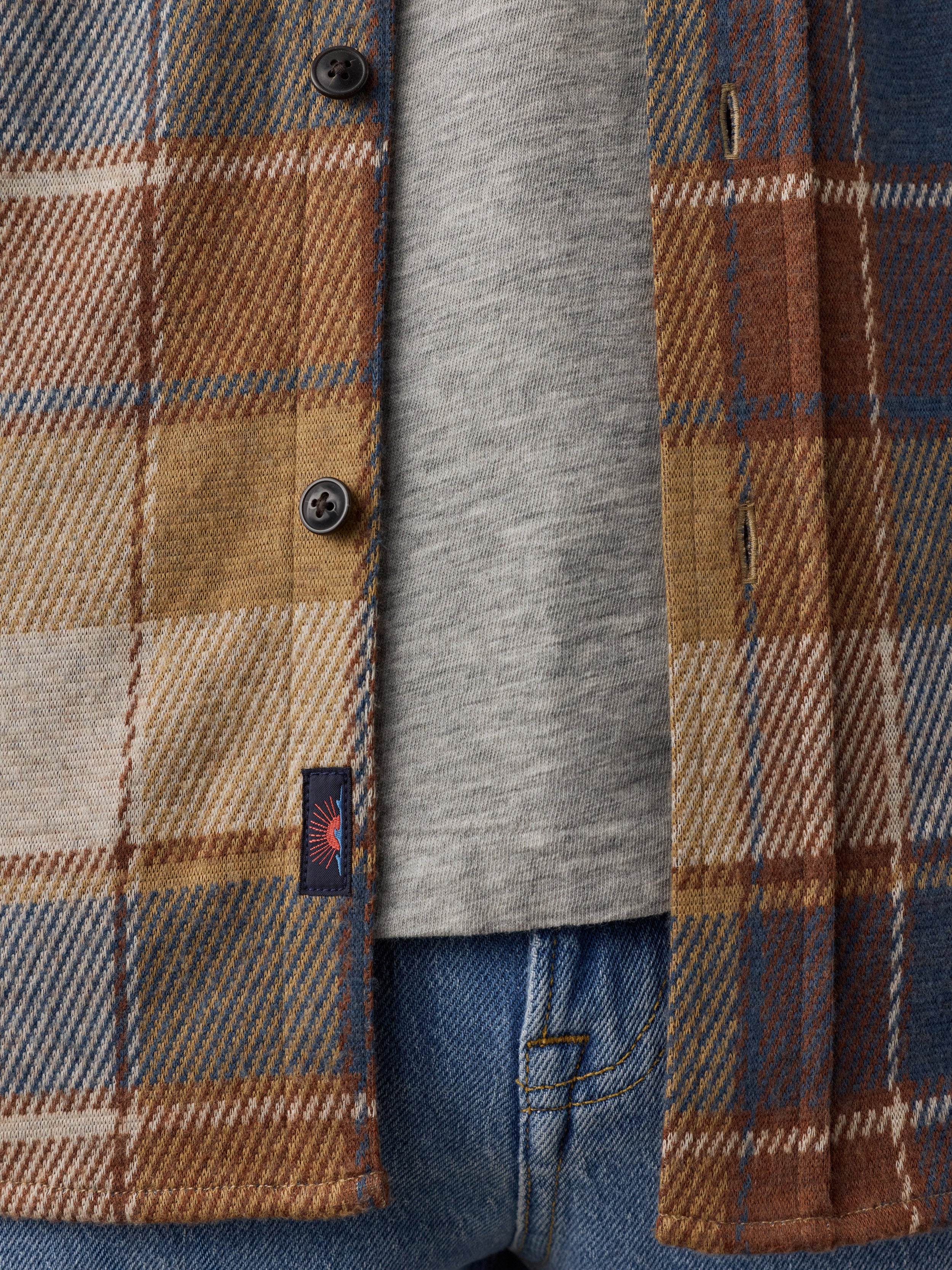 Close-up of a plaid overshirt in tan, brown, and blue tones layered over a heather gray T-shirt and blue jeans, with a small visible Faherty brand tab on the shirt.