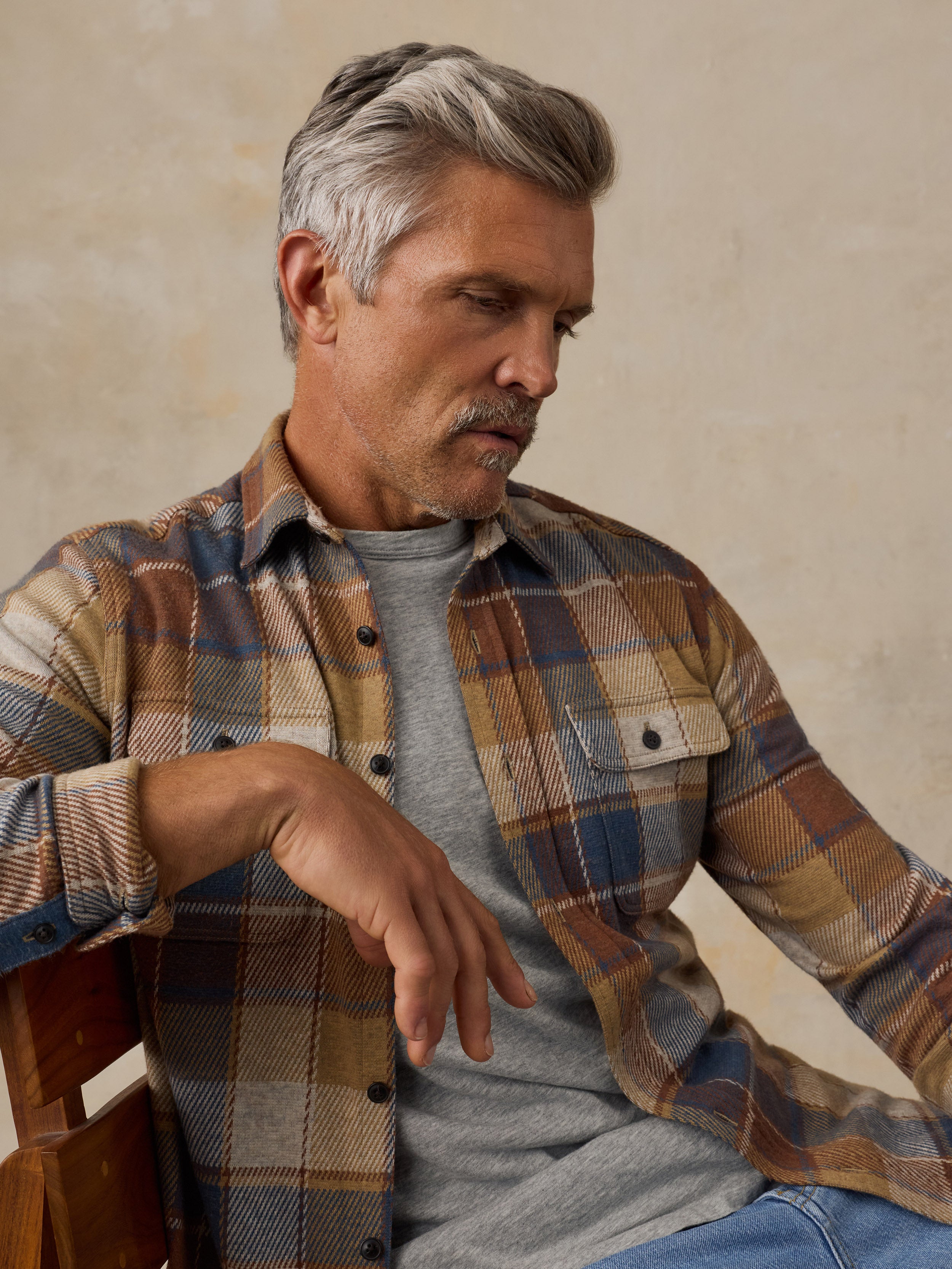 A man with gray hair and a mustache wearing a brown, beige, and blue plaid flannel shirt over a gray t-shirt, sitting on a wooden chair against a textured beige wall.