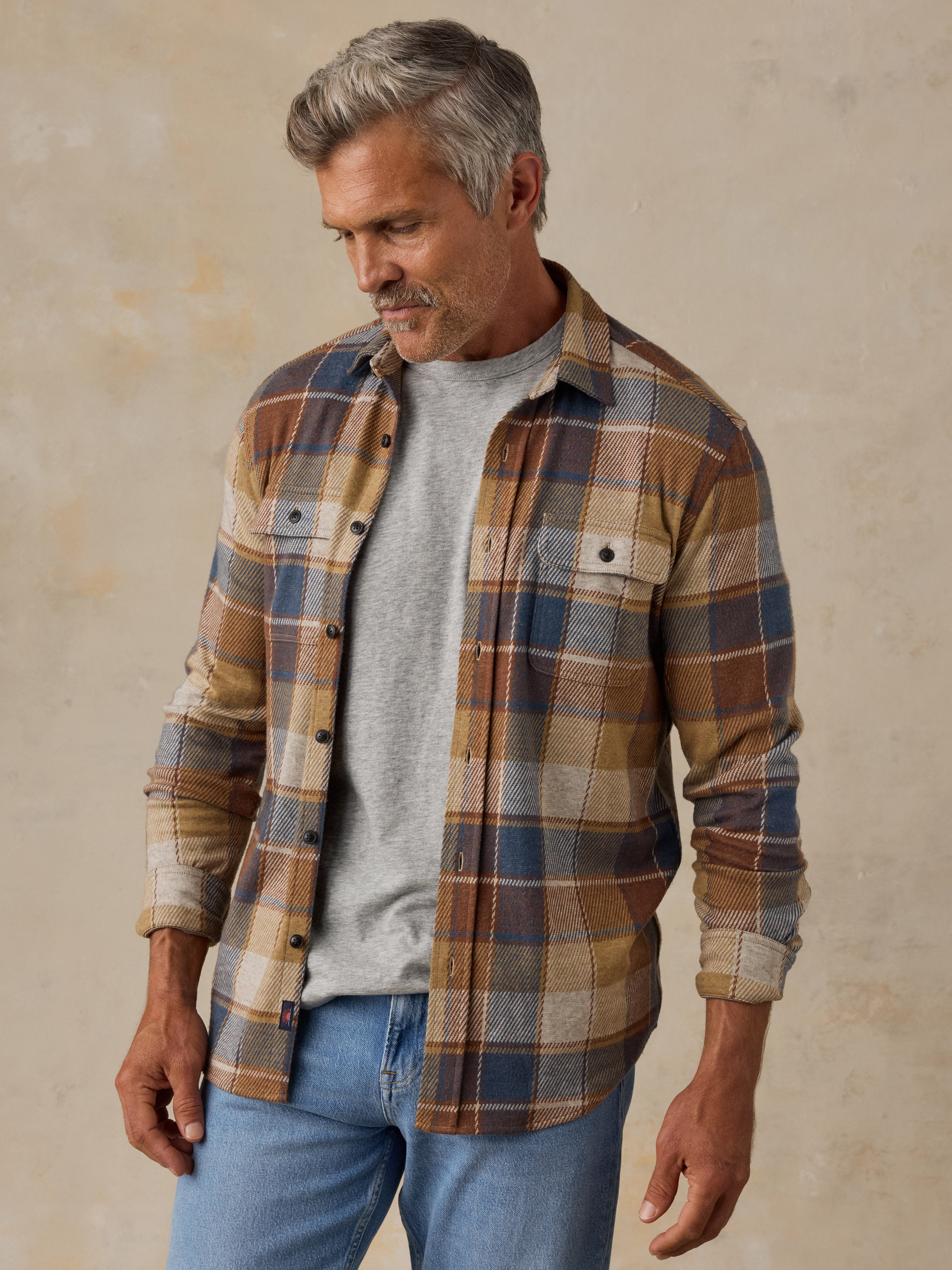 A man wearing a brown, beige, and blue plaid button-up flannel shirt over a gray t-shirt and blue jeans, standing against a textured beige wall.