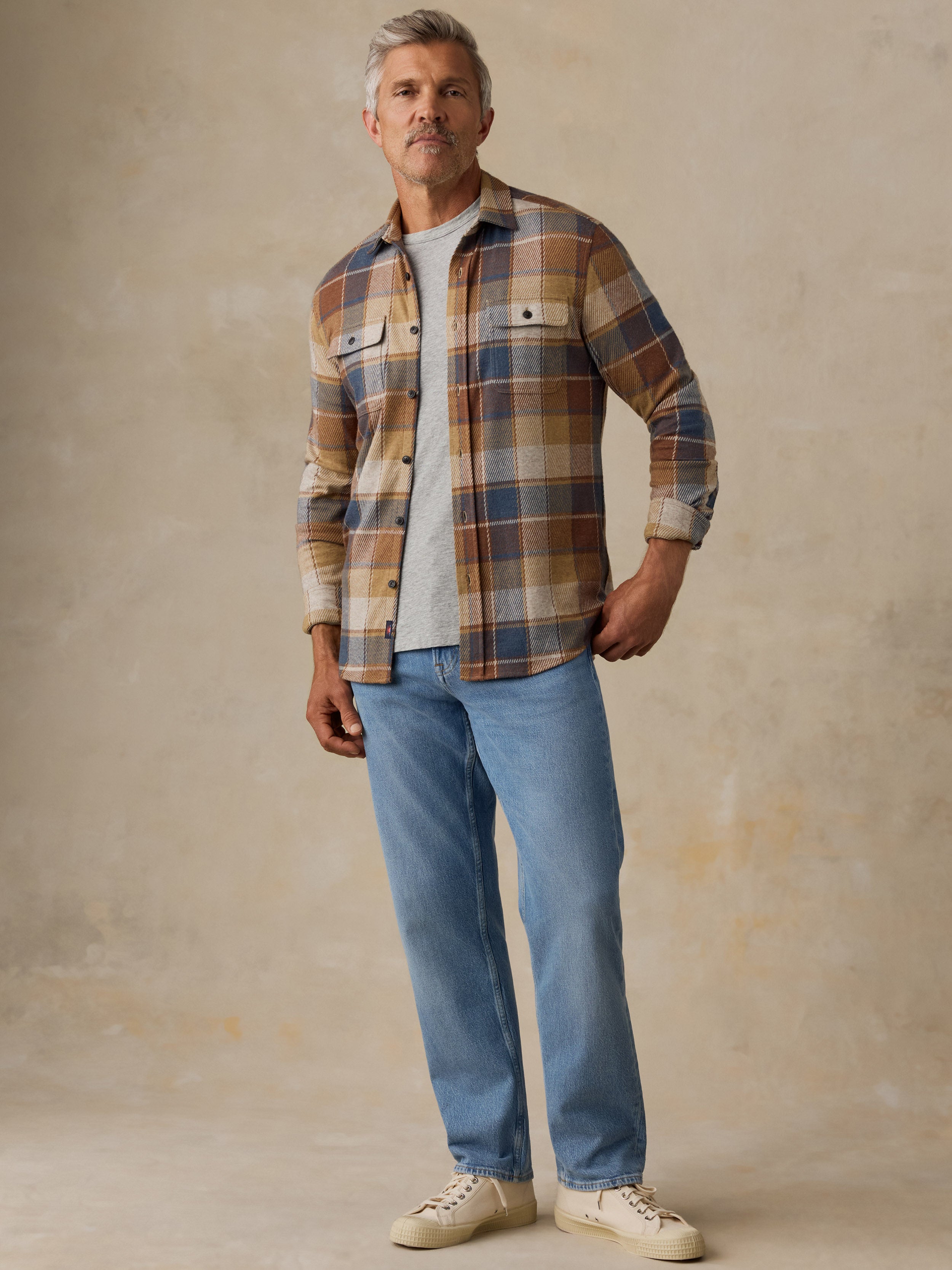 A man modeling a long-sleeve plaid button-up shirt in brown, beige, and blue tones, worn open over a white t-shirt with light blue jeans and cream sneakers, set against a textured beige studio backdrop.