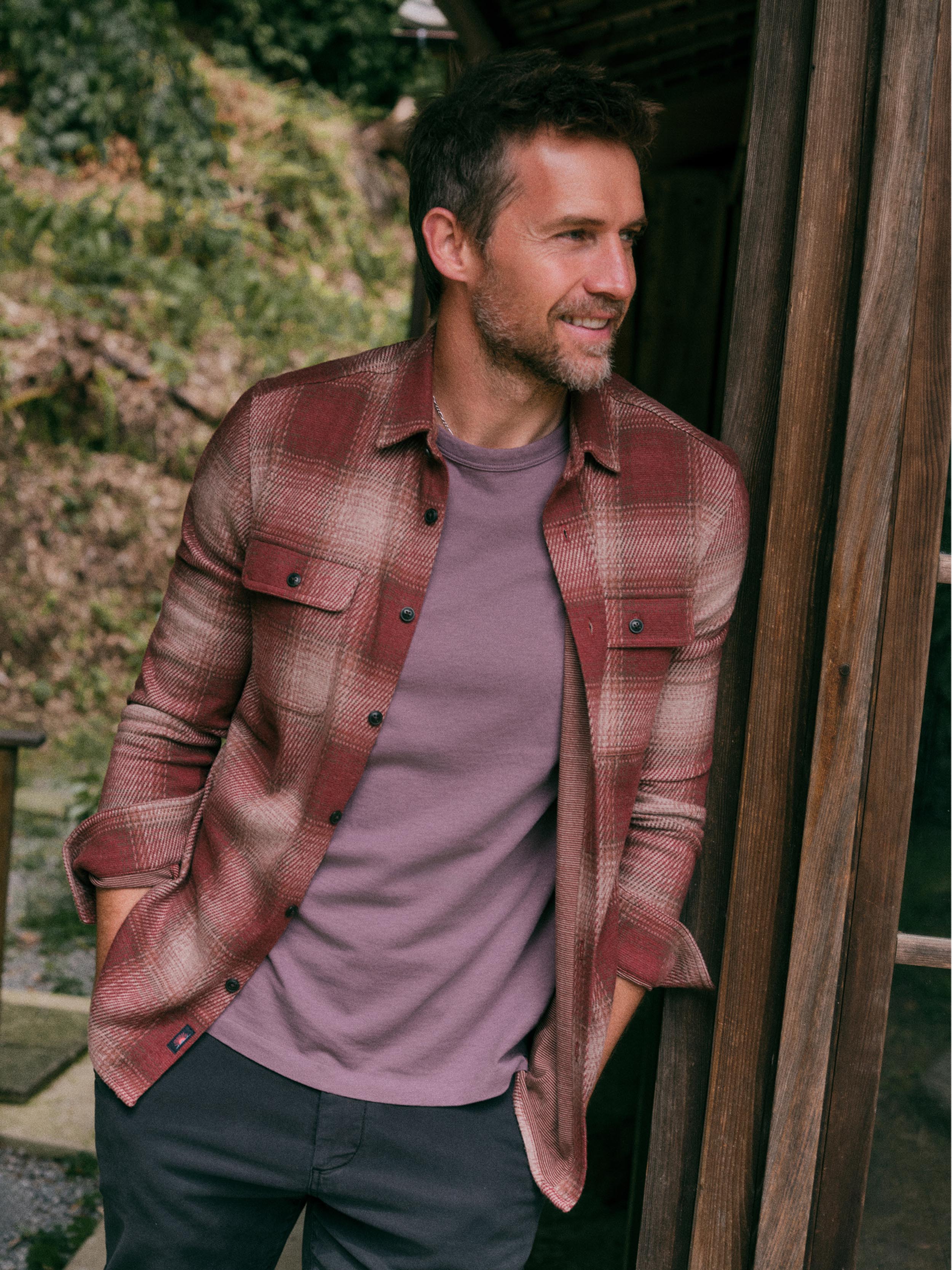 A man wearing a rust red and brown plaid flannel shirt over a mauve-purple t-shirt, standing outdoors next to a wooden structure.