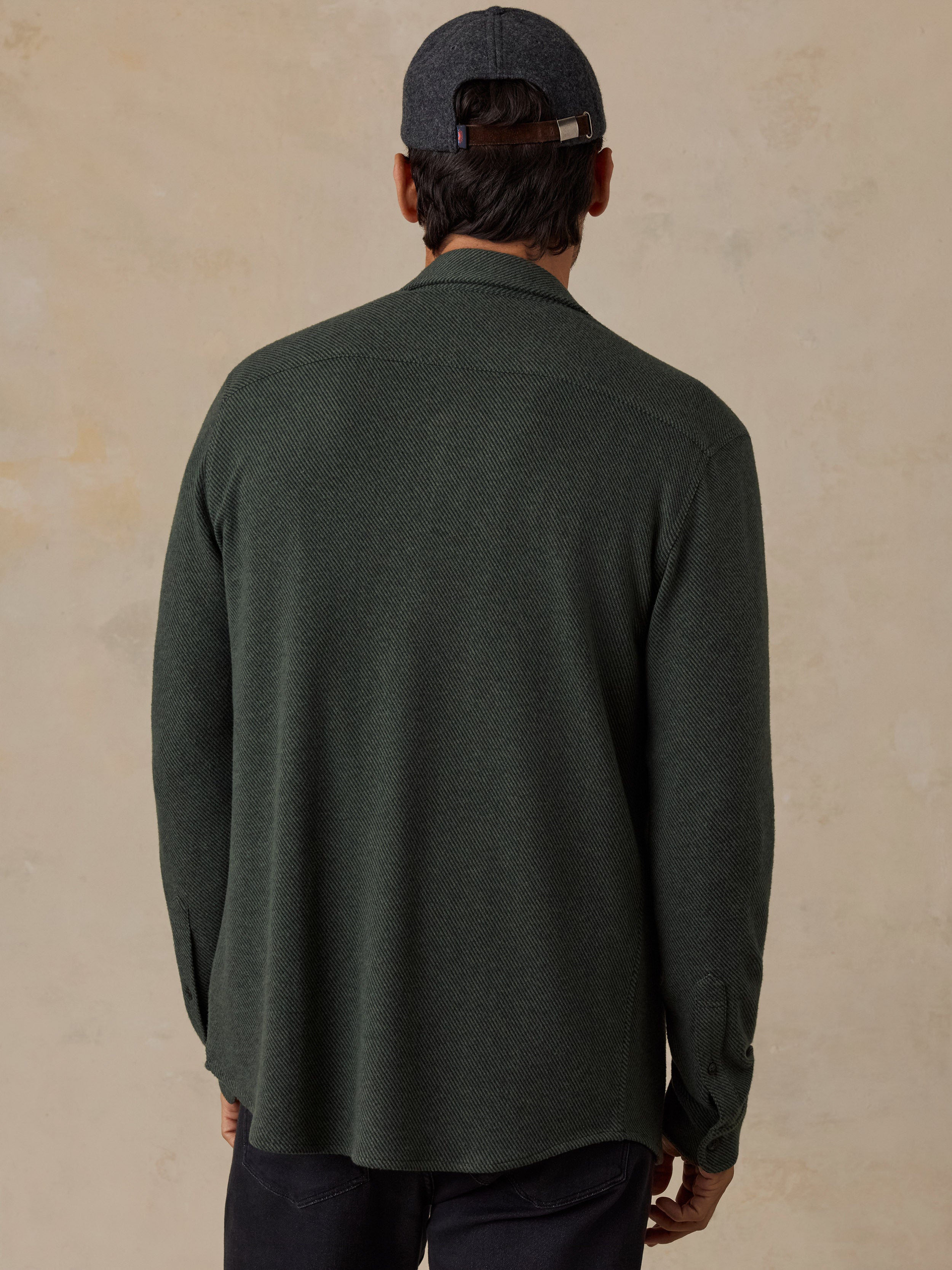 A man viewed from behind wearing a dark olive green long-sleeve knit shirt, a dark gray cap, and dark pants, standing against a textured beige wall.