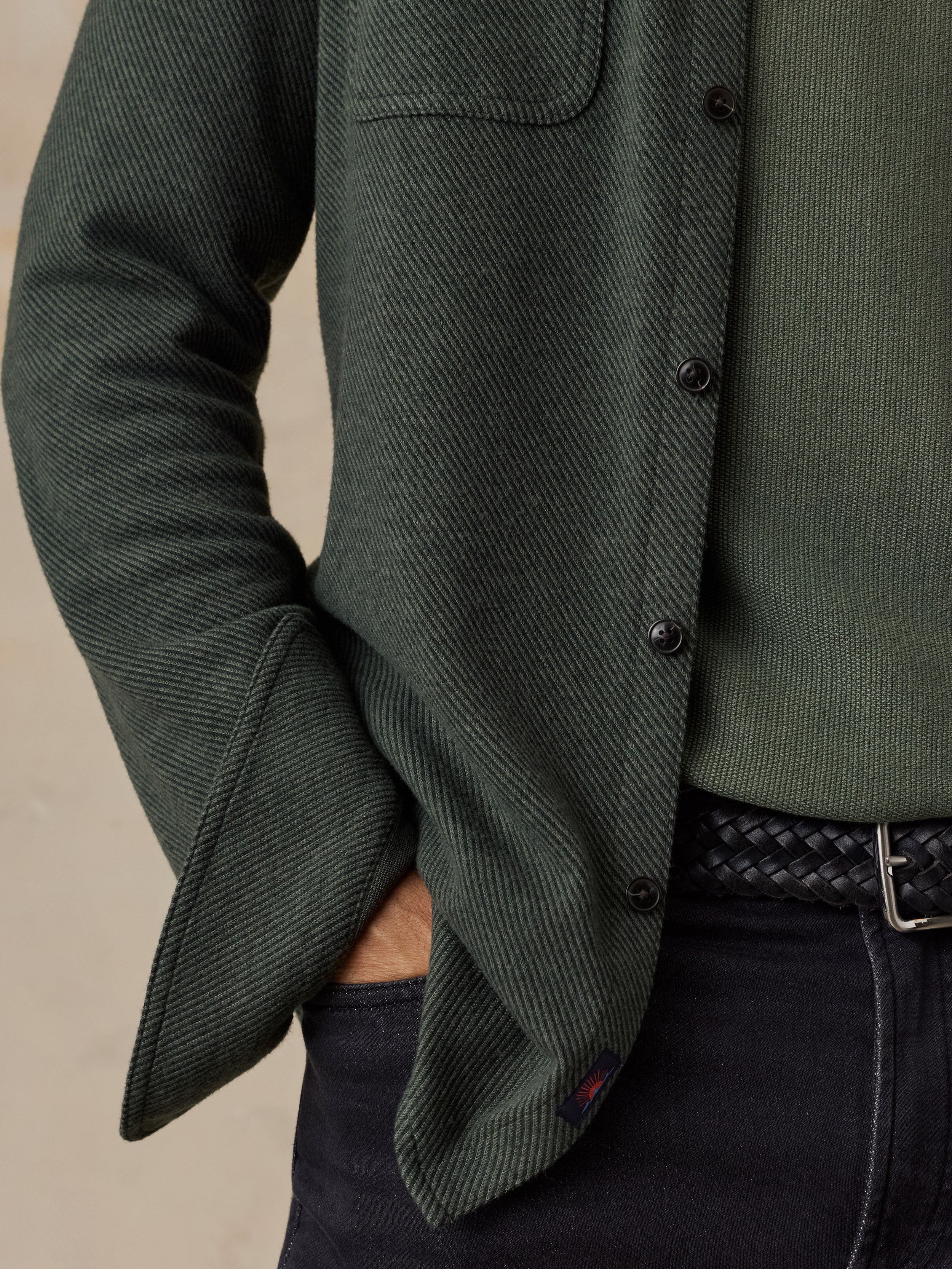 Close-up of a man wearing a dark green textured overshirt over an olive green knit sweater, paired with dark jeans and a black braided belt.