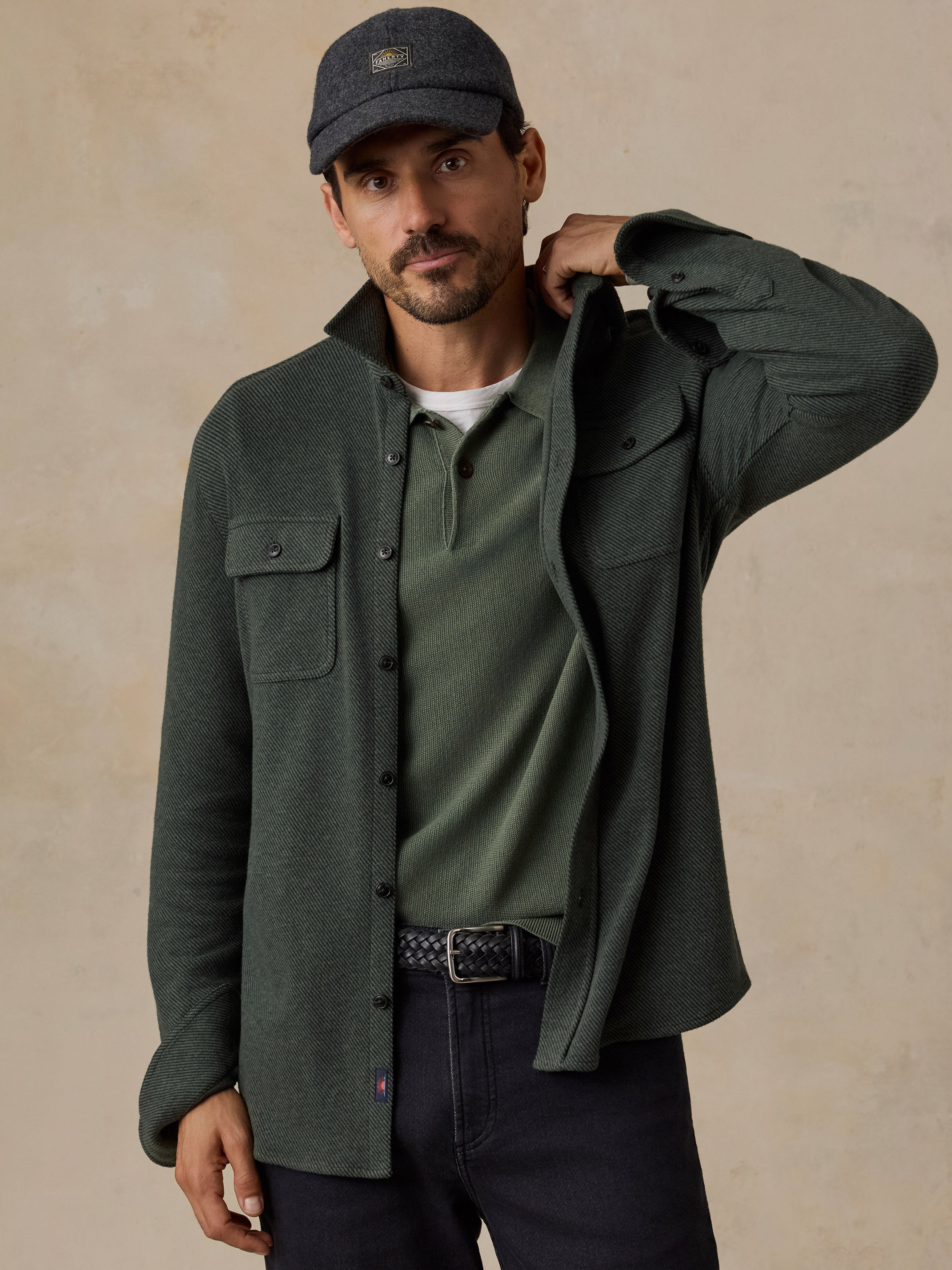 A man wearing a dark olive green button-up overshirt over a matching green henley shirt, dark jeans, and a dark gray baseball cap, posed against a textured beige background.