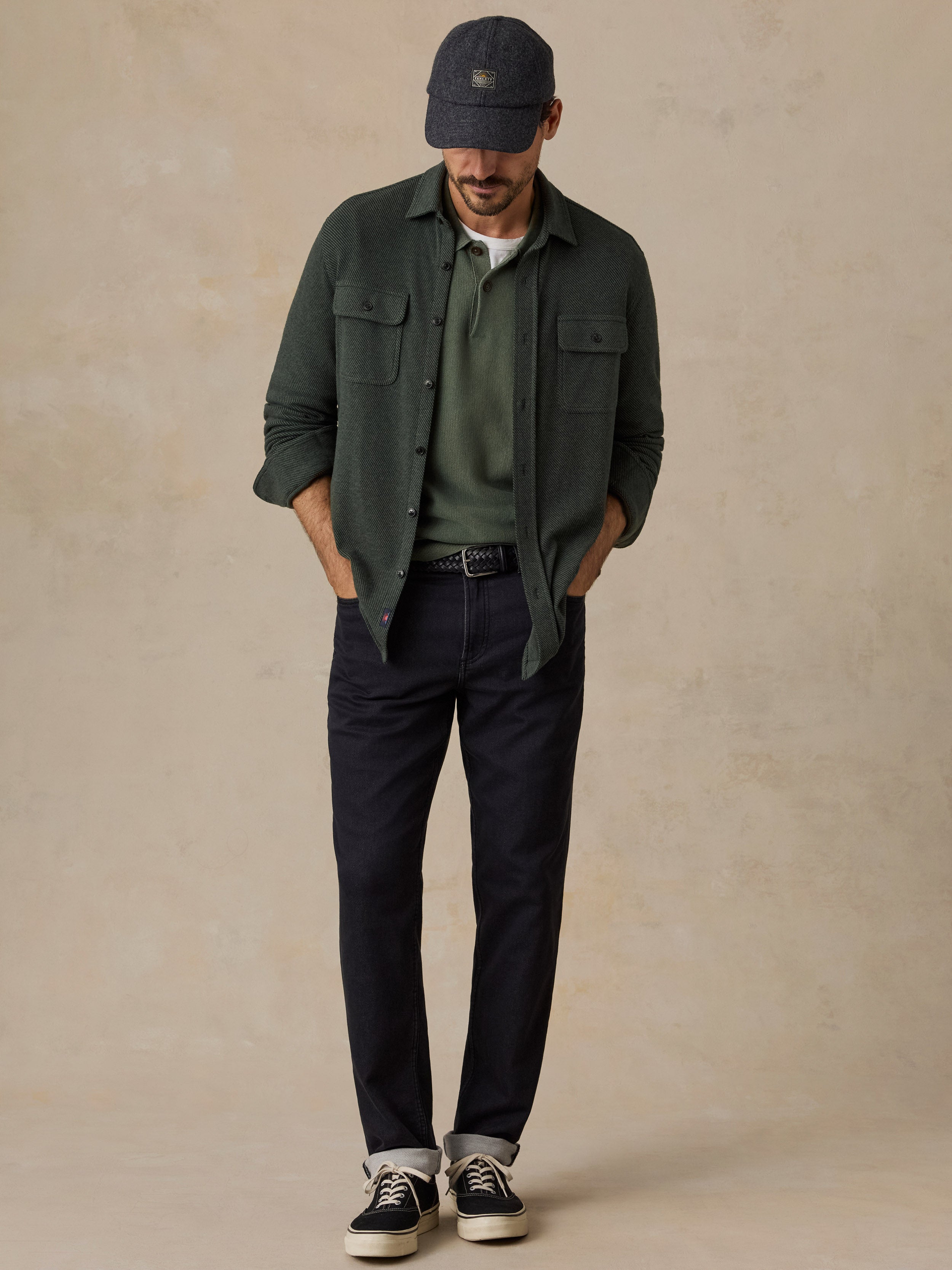 A man wearing a dark green overshirt over an olive green t-shirt, dark navy pants, black sneakers, and a dark cap, standing against a textured beige studio backdrop.