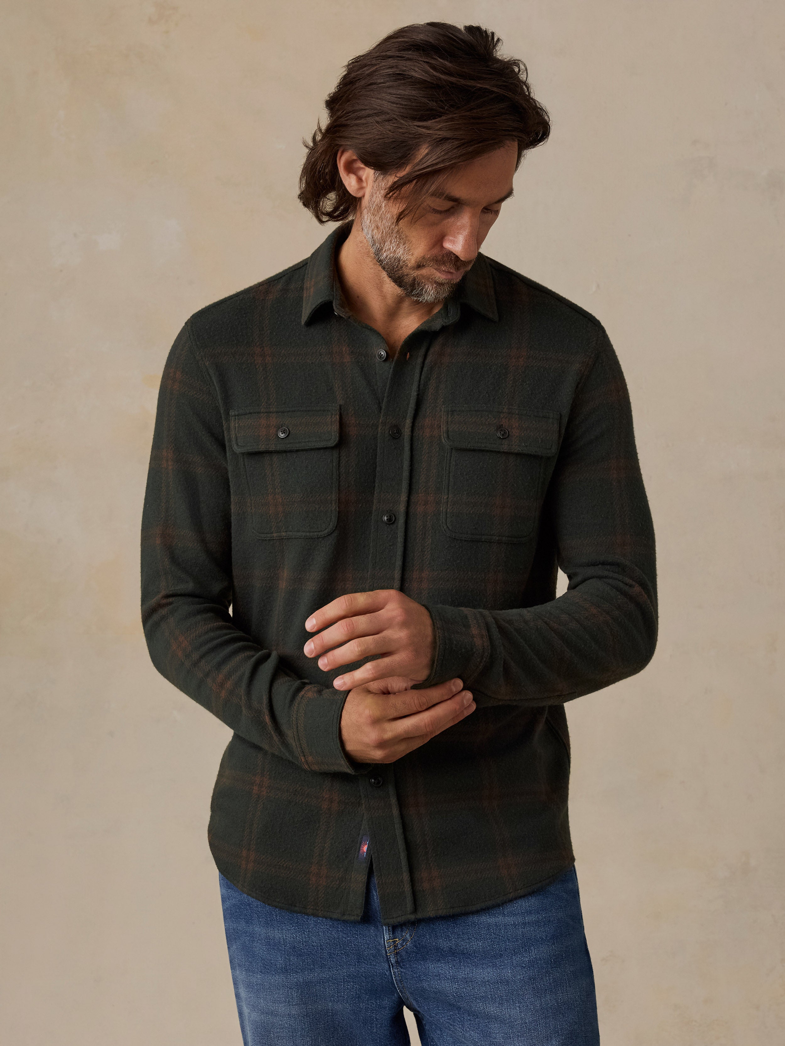 A man wearing a dark green long-sleeve plaid button-up shirt with two chest pockets and blue jeans, standing against a textured beige background.