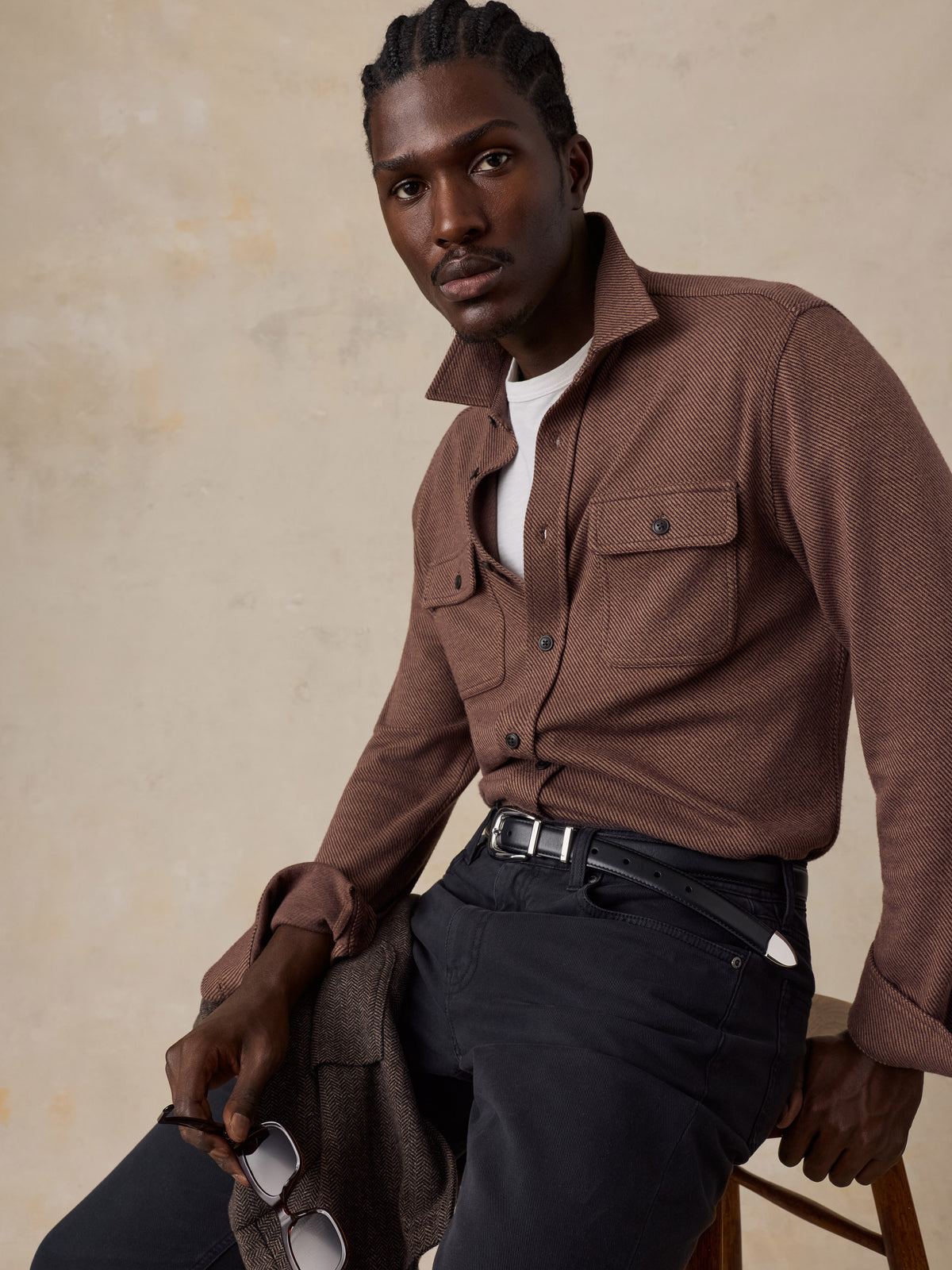 A man sitting on a chair wearing a brown shirt over a white tee and black jeans , image 2 of 5