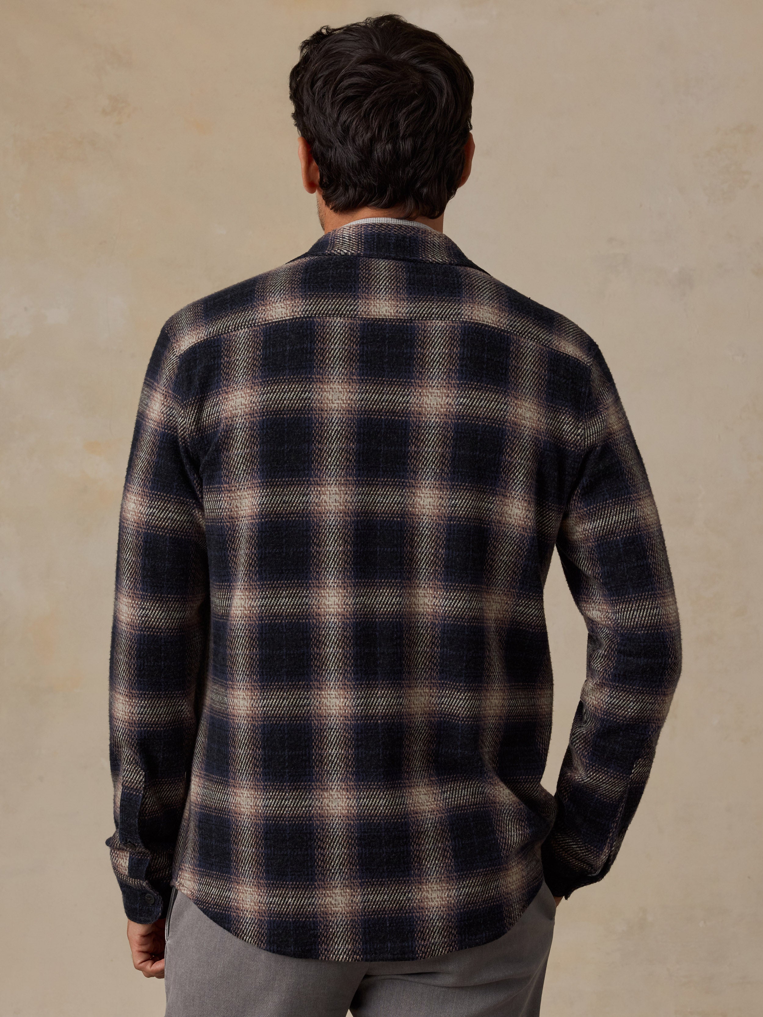 A man viewed from behind wearing a dark navy and beige plaid flannel shirt, paired with gray trousers, against a textured beige background.