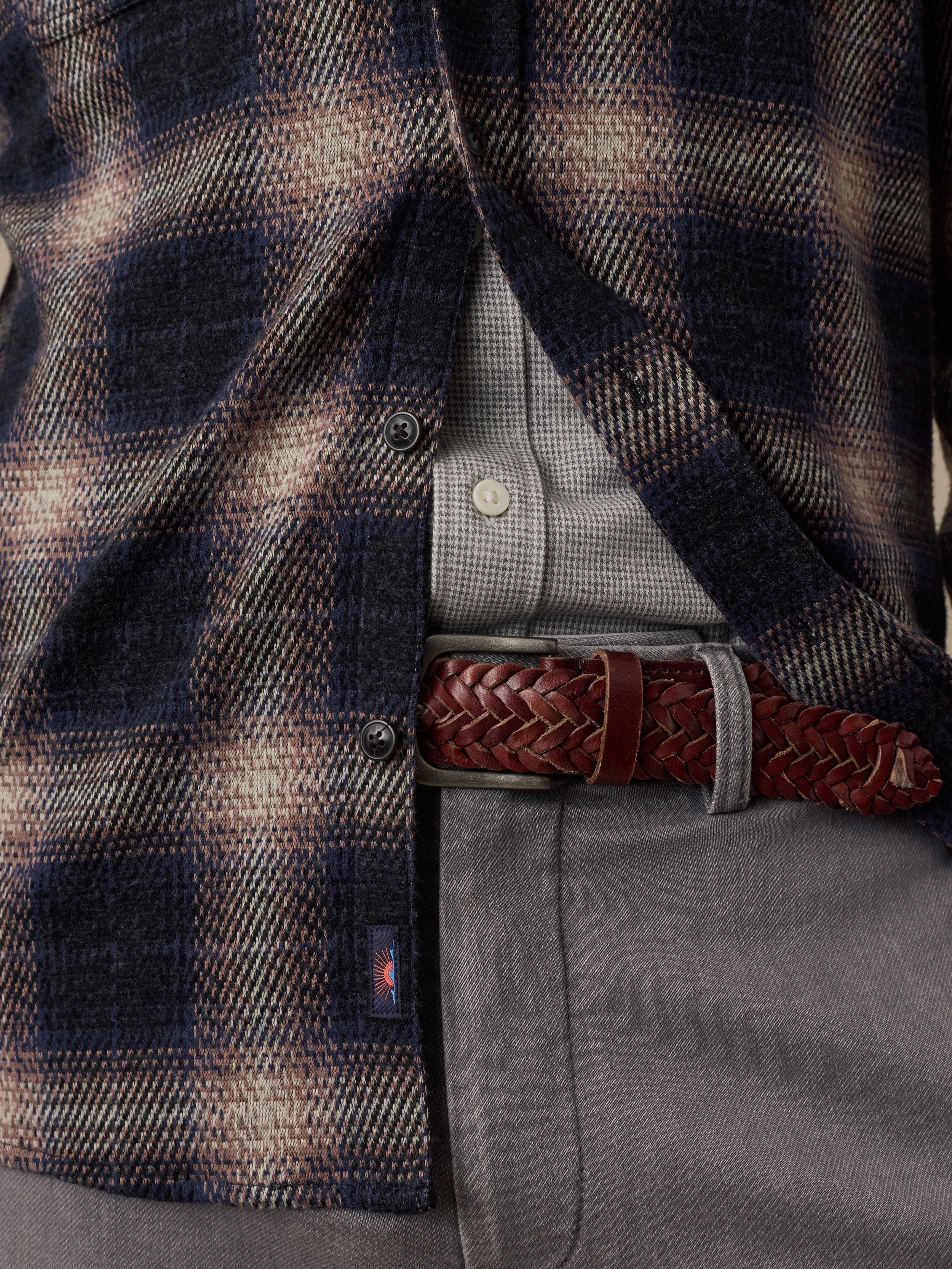 Close-up of a man wearing a navy and beige plaid overshirt over a gray button-up shirt, with a brown braided belt and gray trousers.