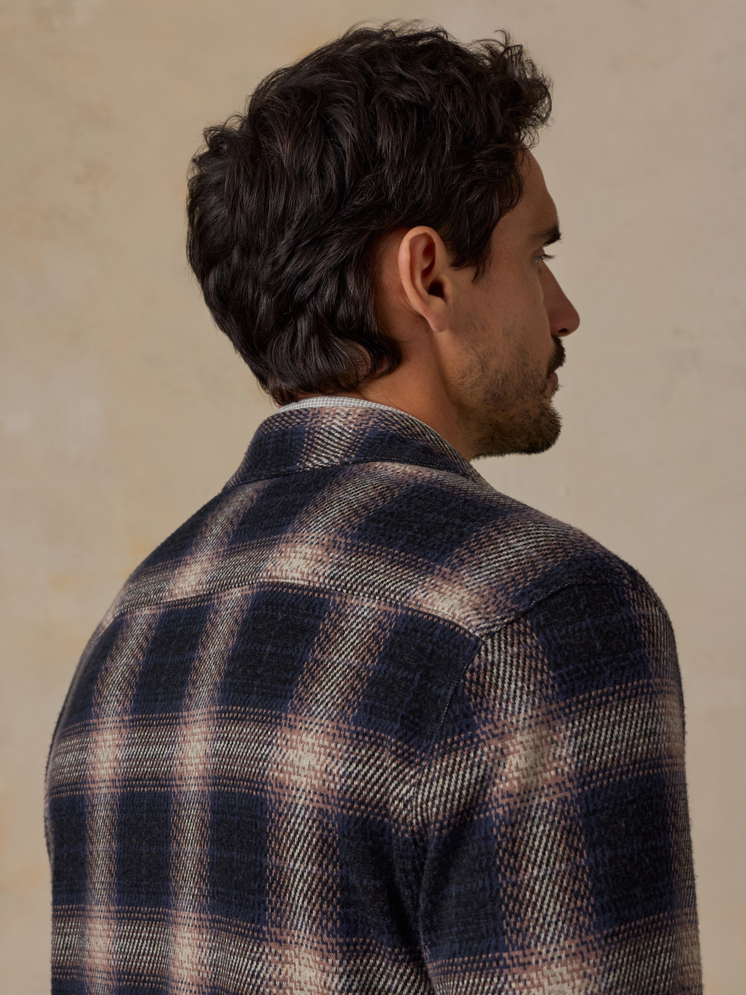 A man shown from the back and side profile wearing a dark navy and beige plaid wool-blend shirt jacket against a textured beige background.