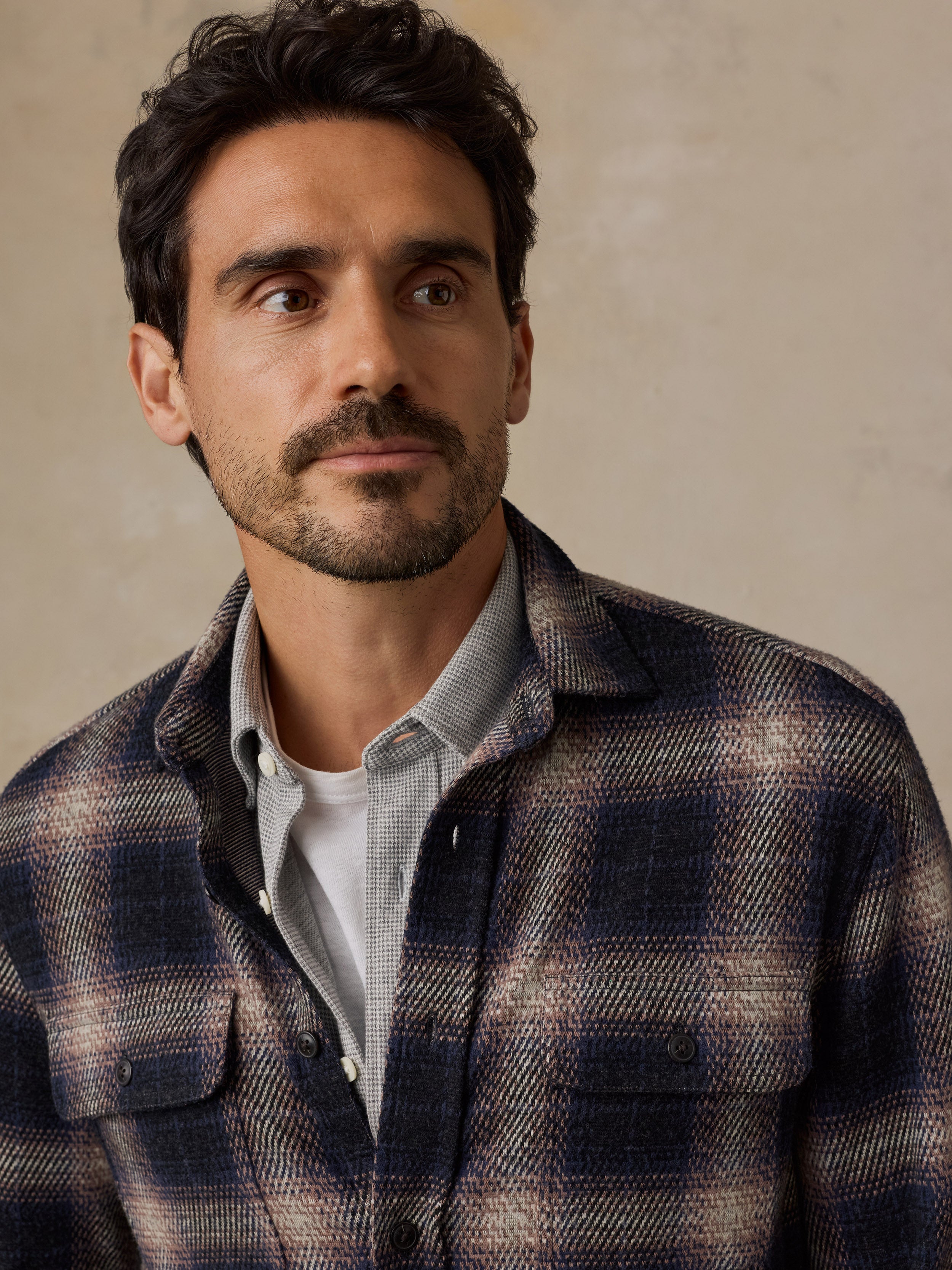 A man wearing a dark navy and brown plaid overshirt over a light gray button-down shirt and white t-shirt, photographed against a textured beige background.