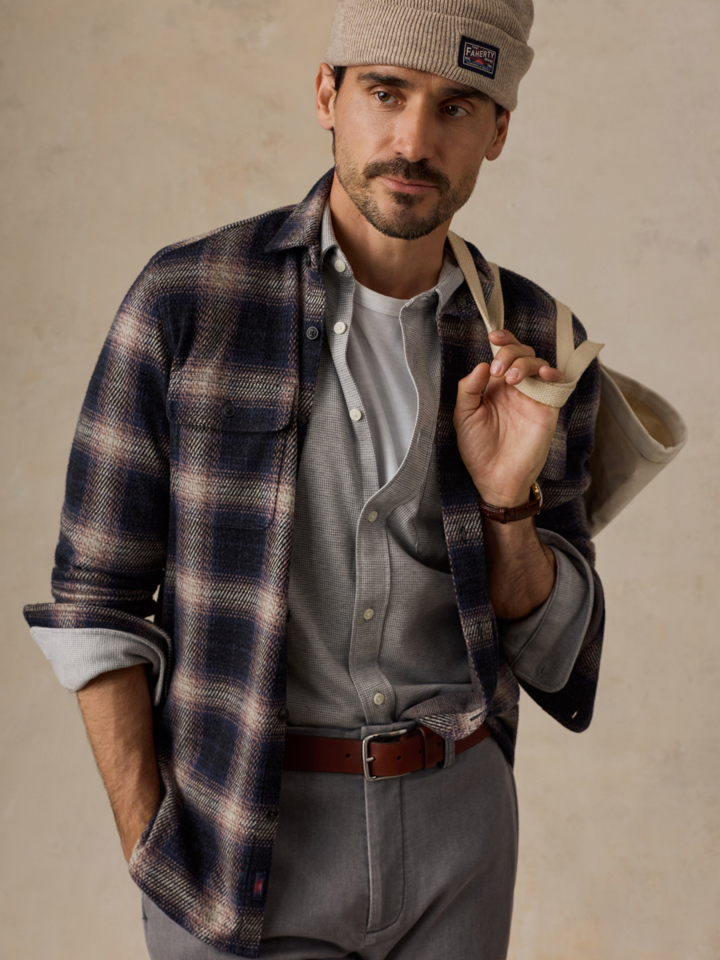 A man wearing a navy and beige plaid flannel shirt over a gray button-up shirt, paired with gray trousers, a brown leather belt, a beige knit beanie, and carrying a canvas tote bag.