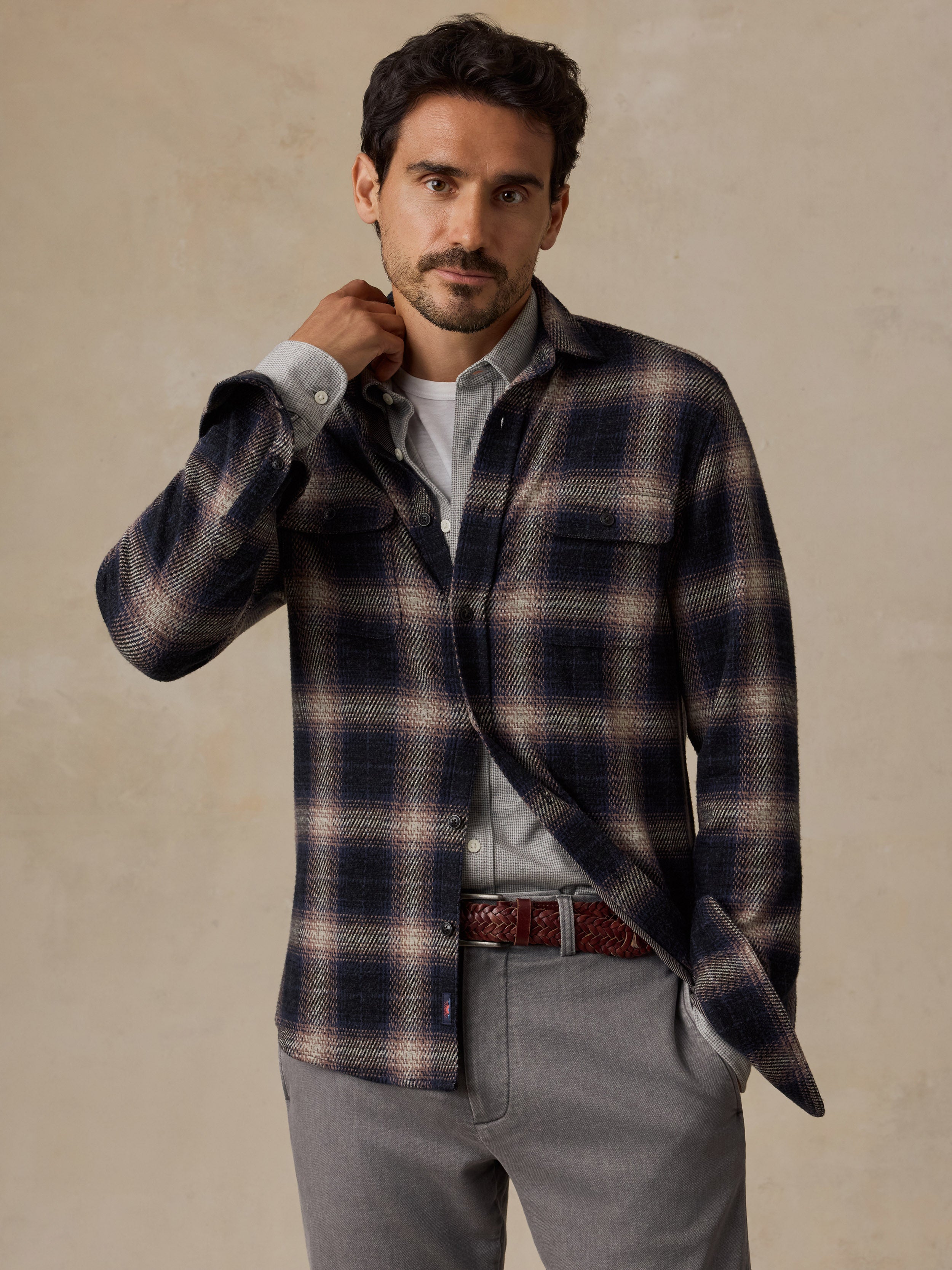 A man wearing a navy and beige plaid overshirt over a white shirt, paired with gray trousers and a brown belt, standing against a textured beige background.