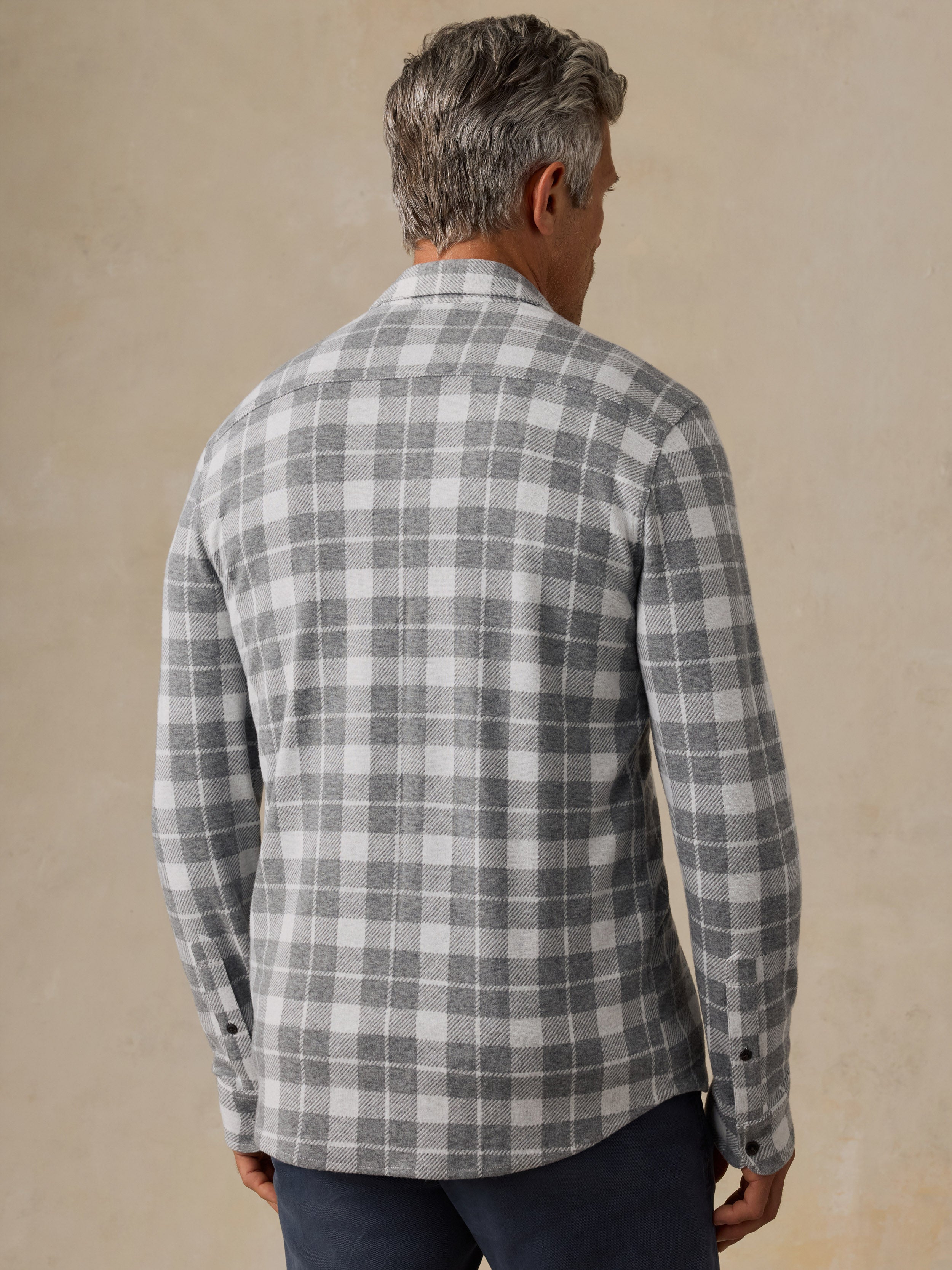 A man viewed from behind wearing a gray and white plaid button-down shirt and dark trousers, standing against a textured beige wall.