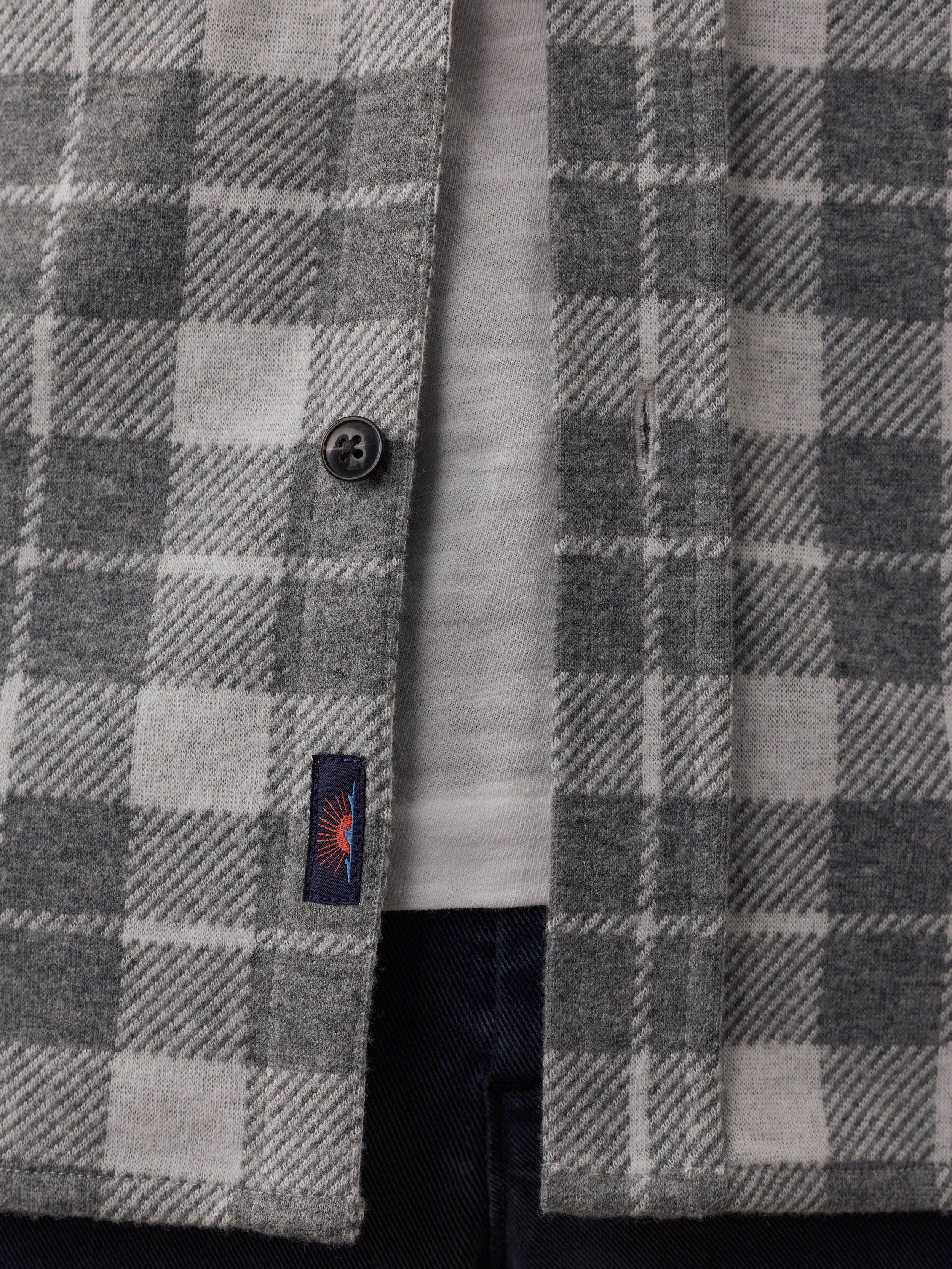 Close-up of a gray and white plaid button-up shirt jacket worn over a light gray t-shirt, showing a small Faherty brand tag with a sun and waves logo.