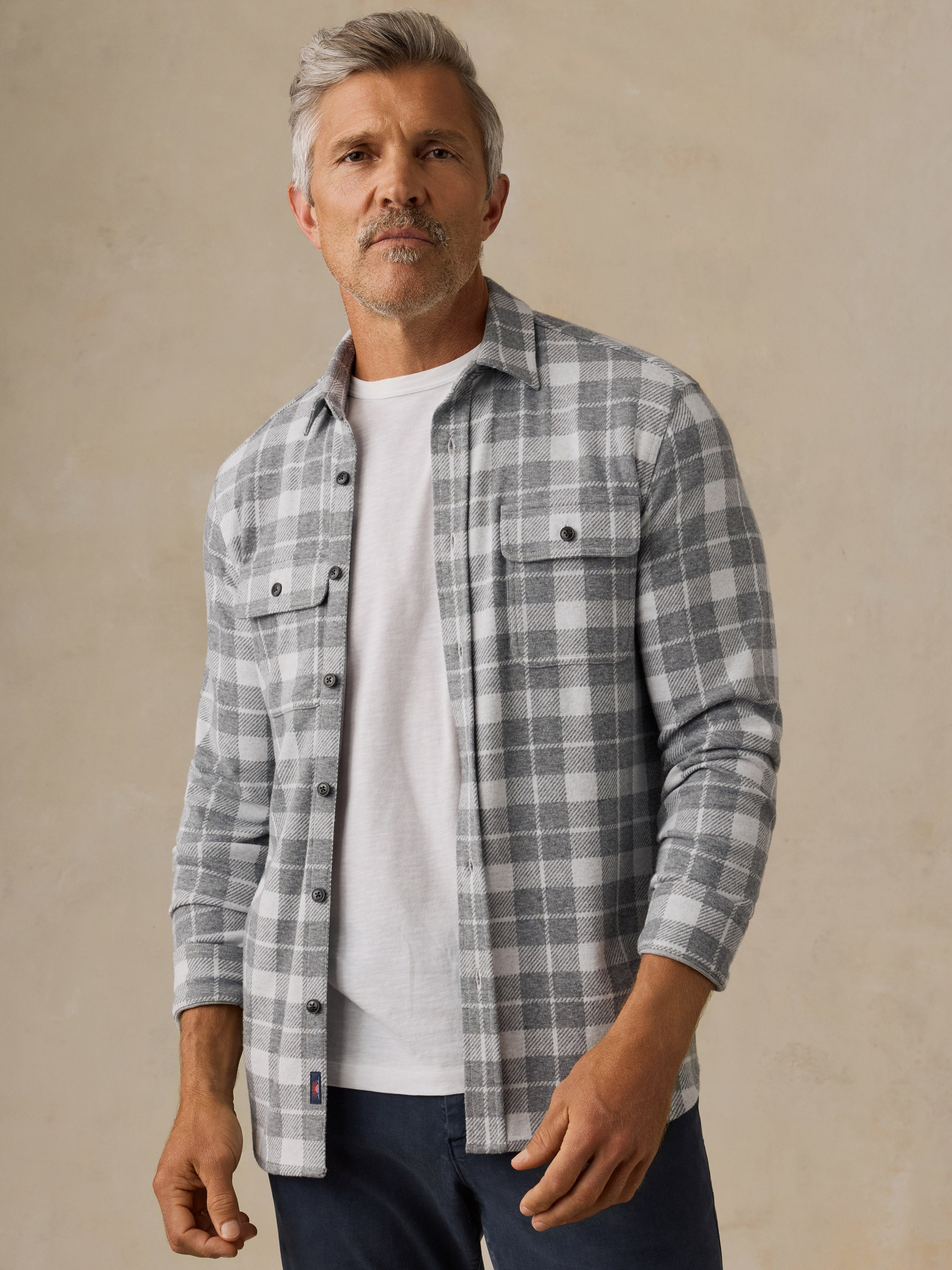 A man wearing a gray and white plaid button-down flannel shirt over a white t-shirt and navy pants, standing against a textured beige background.