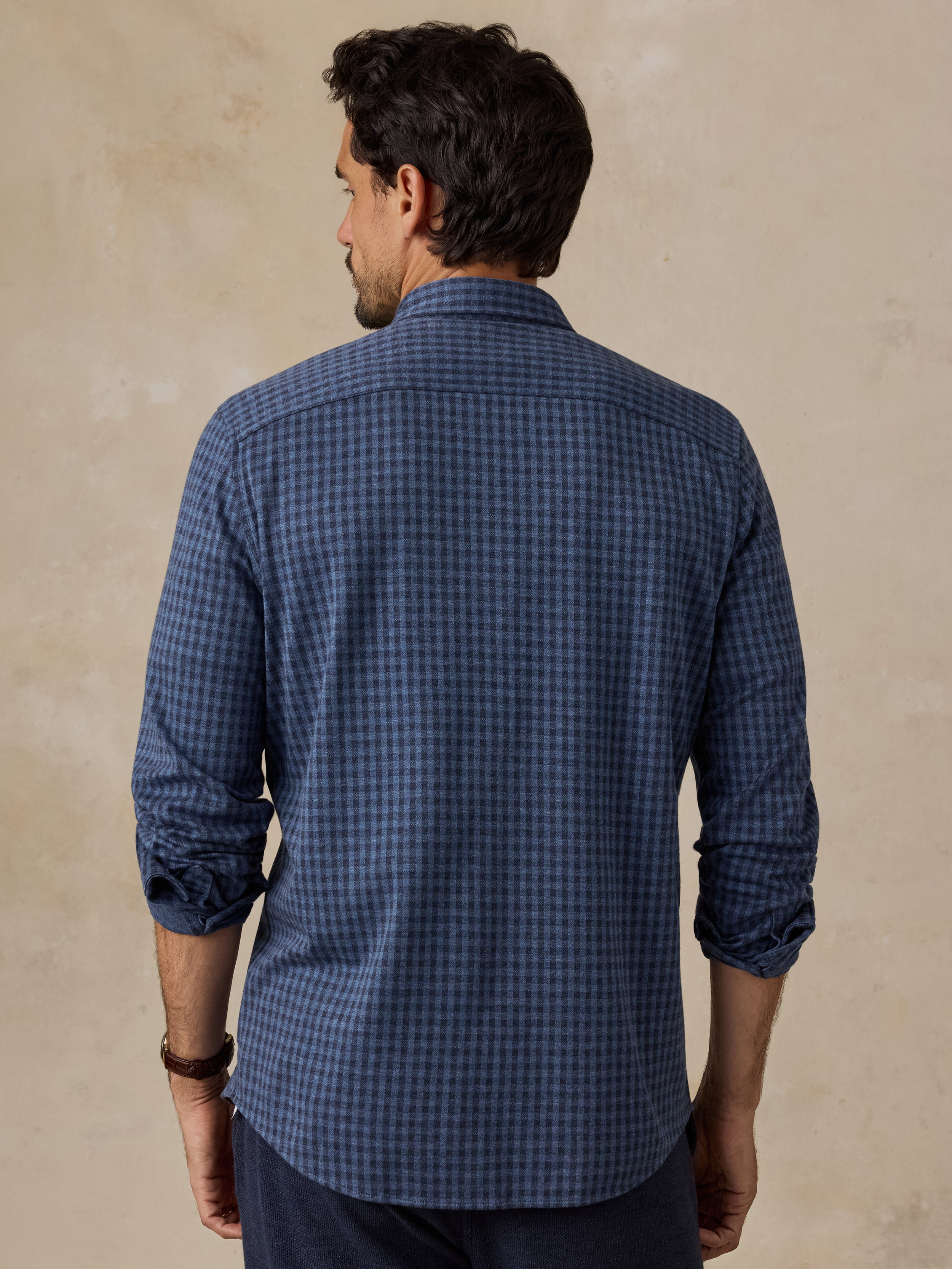 A man viewed from the back wearing a blue and black checkered long-sleeve button-down shirt, set against a textured beige background.