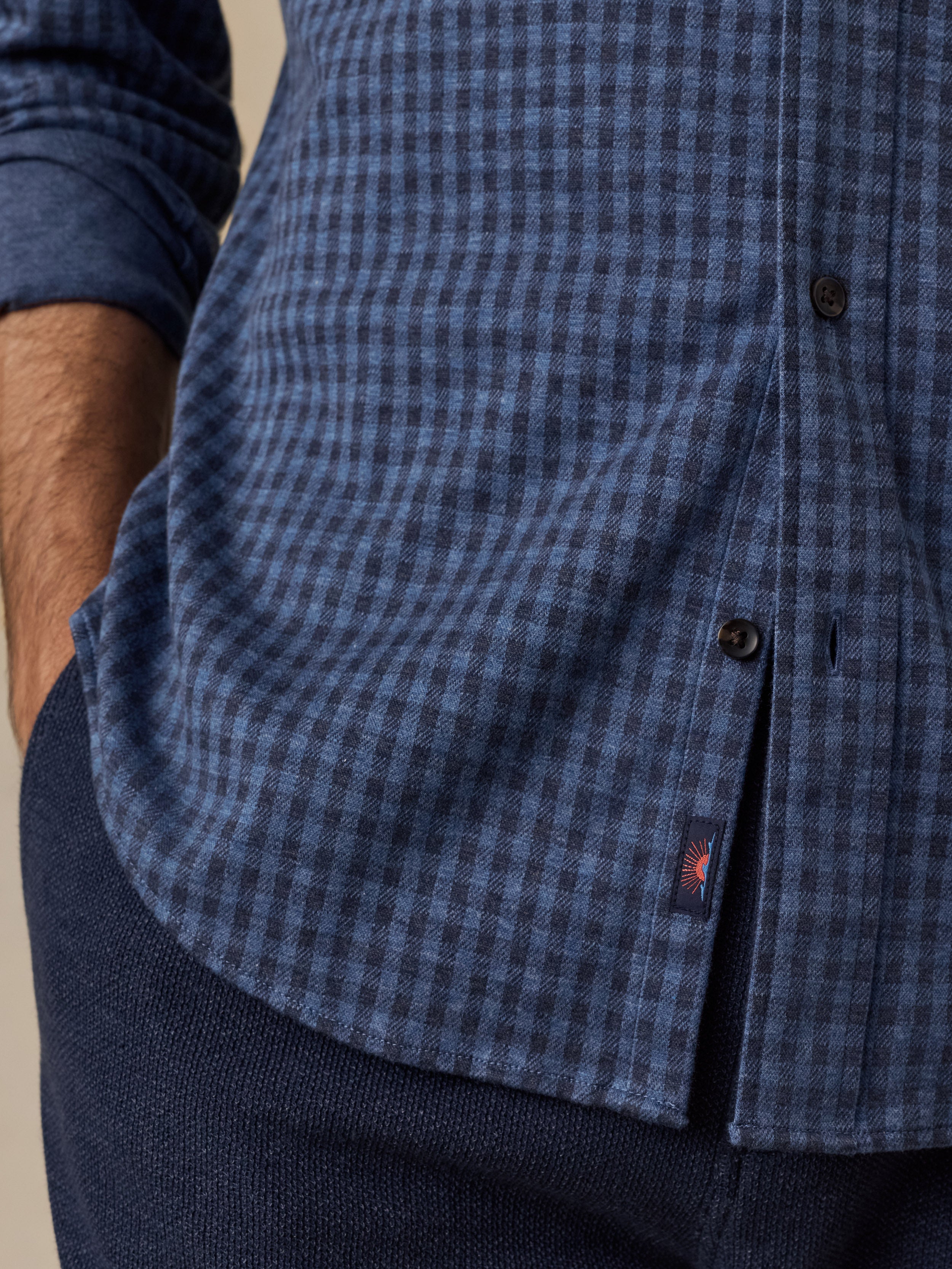 Close-up of a man wearing a blue and navy checkered button-down shirt with dark navy trousers, showing the fabric texture and a small red Faherty brand tab.